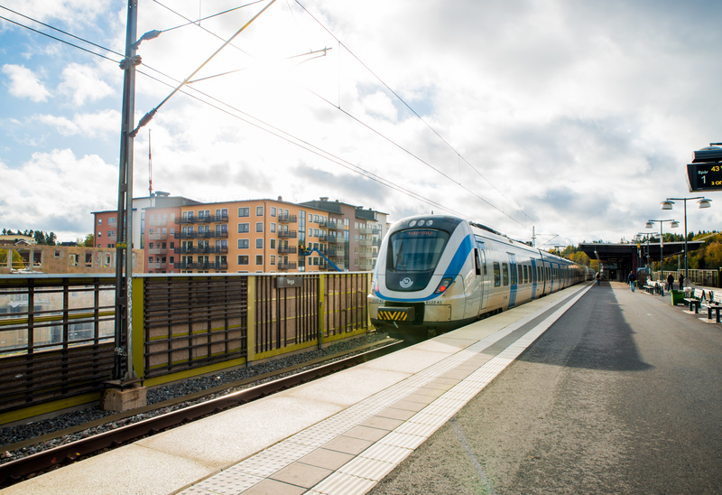 Pendeltåg rullar in på perrongen med flerfamiljshus i bakgrunden.