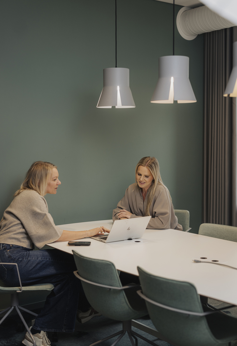 Två personer sitter mitt emot varandra vid ett vitt konferensbord i ett modernt mötesrum och pratar, medan den ena arbetar på en öppen bärbar dator. Rummet har gröna väggar, fyra gröna stolar och tre vita pendellampor som hänger ovanför bordet.