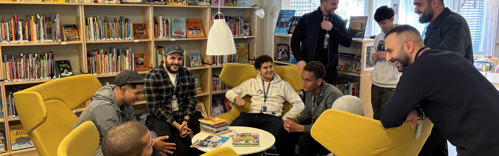 10 killar i samtal i ett bibliotek. Fem av dem sitter i på gula stolar runt ett runt bord. Resten står upp. I bakgrunden syns bokhyllor fyllda med böcker