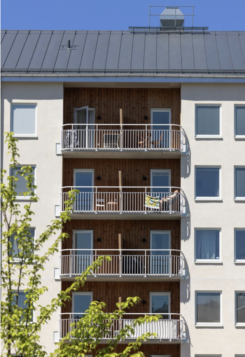 Fasad av ett modernt flerbostadshus med vita väggar och en mittsektion i trä, där flera balkonger med vita räcken ligger ovanpå varandra; på balkongerna syns utemöbler och en person som solar, och i förgrunden skymtar gröna träd mot en klarblå himmel.