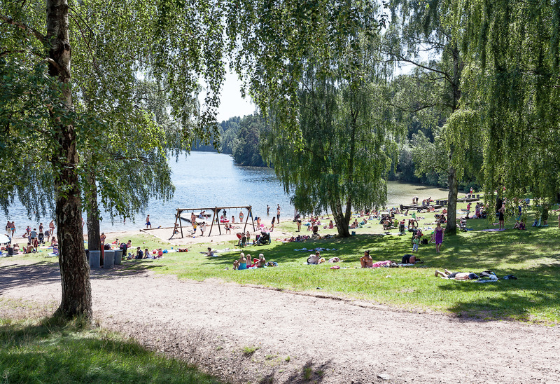 En solig dag vid en sjö med en sandstrand där människor badar och solar. Gräsmattan är fylld med folk som kopplar av under stora träd. En lekplats med gungor syns i bakgrunden. Området är grönt och lummigt.