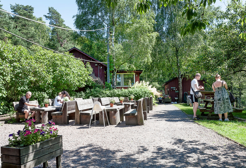En utomhusservering med träbänkar och bord omgiven av grönska. Människor sitter och äter eller står och pratar. I bakgrunden syns röda träbyggnader och träd. Det är en solig dag.
