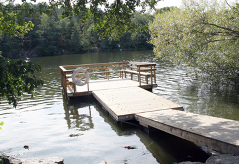 En brygga i vatten inringat av grönskande träd. En träbrygga som sträcker sig ut i en lugn sjö, omgiven av grönskande träd. En livboj hänger på bryggan, och vattnet är stilla under en klar himmel. Stämningen är fridfull och naturskön.