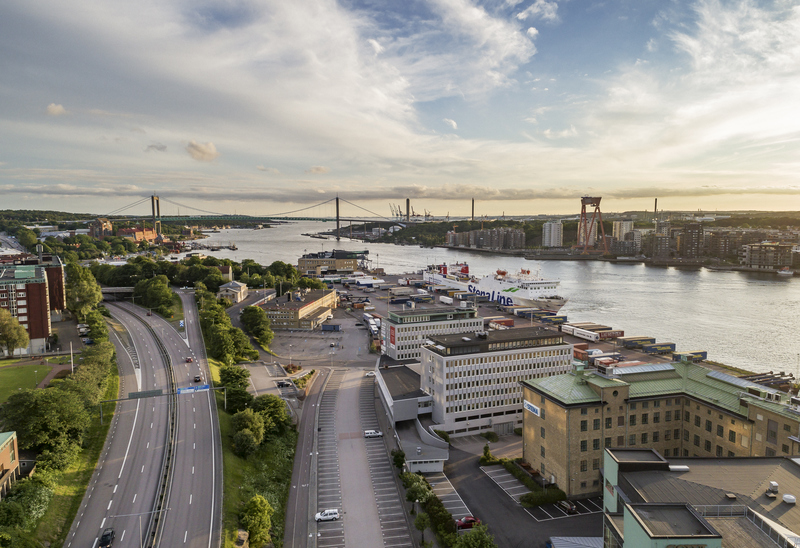 Överblicksbild på Göteborg, Fiskhamnsgatan. Här ser man vägar, fastigheter, älven och Älvsborgsbron.En flygbild över en stad med en bred flod som sträcker sig genom landskapet. Flera broar korsar floden, och hamnområden med båtar syns längs vattnet. Omgivningen består av byggnader och grönområden under en himmel med spridda moln och solnedgångsljus. 
