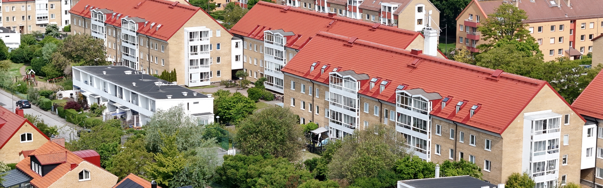 En tätbebyggd bostadsmiljö med flerfamiljshus i tegelfasad och röda tak omgivna av trädgårdar, träd och mindre villor i förgrunden.