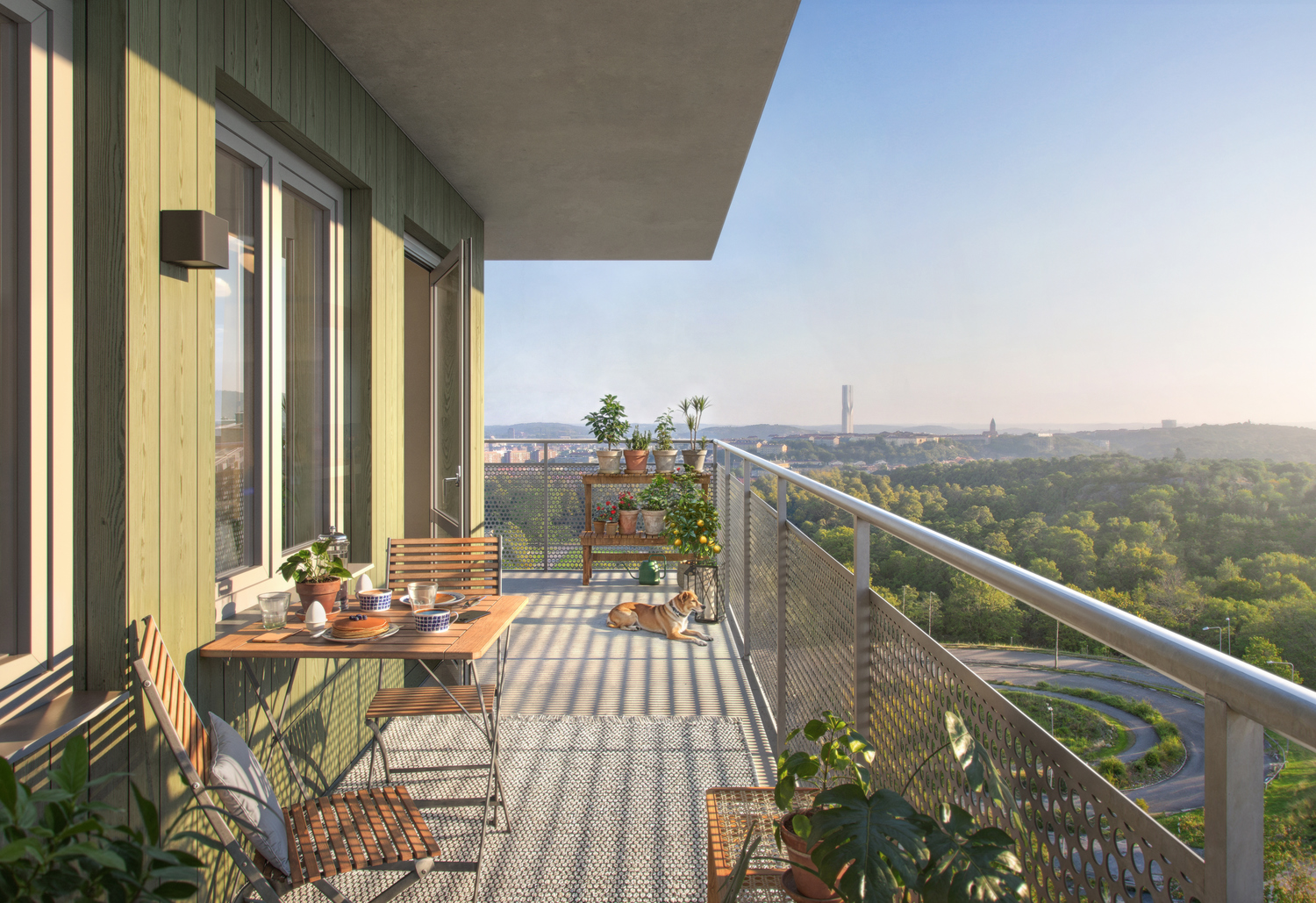 En solig balkong med stora gröna glasdörrar och träbord med stolar. Växter i krukor och rostfritt metallräcke. Utsikt över grönt landskap, träd och en avlägsen stadsvy under en klar blå himmel