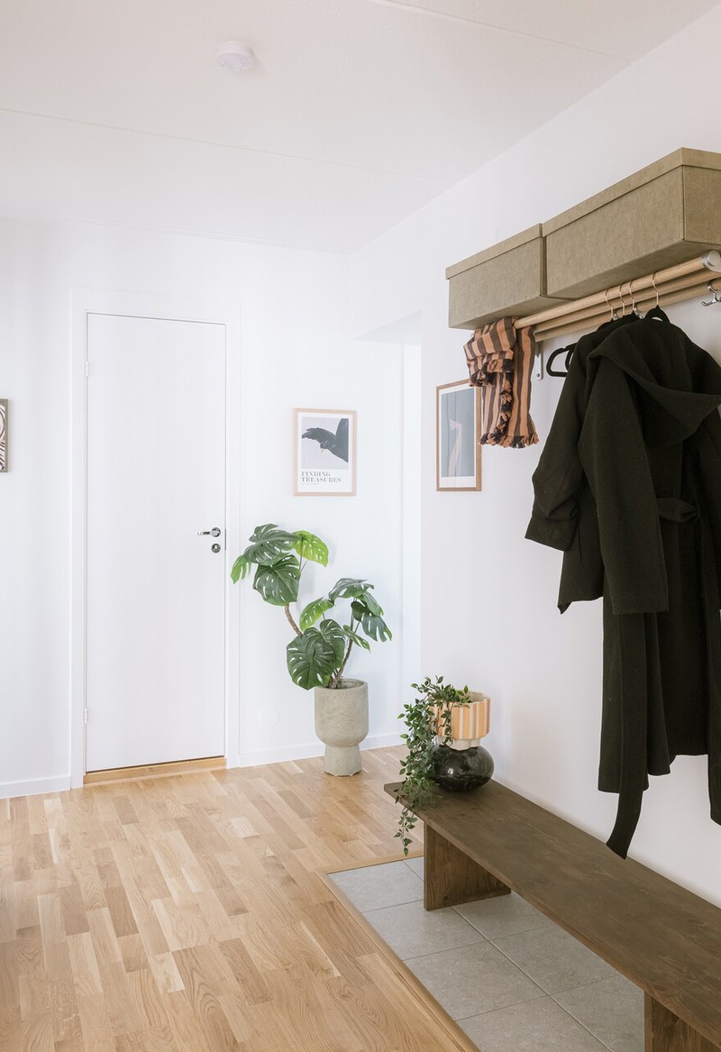 Ljus och rymlig hall med ljust trägolv, vitmålade väggar, en träbänk med växt och ljuslykta, ytterkläder hängande på en hatthylla samt spegel, sittpuff och gröna krukväxter som inredning.