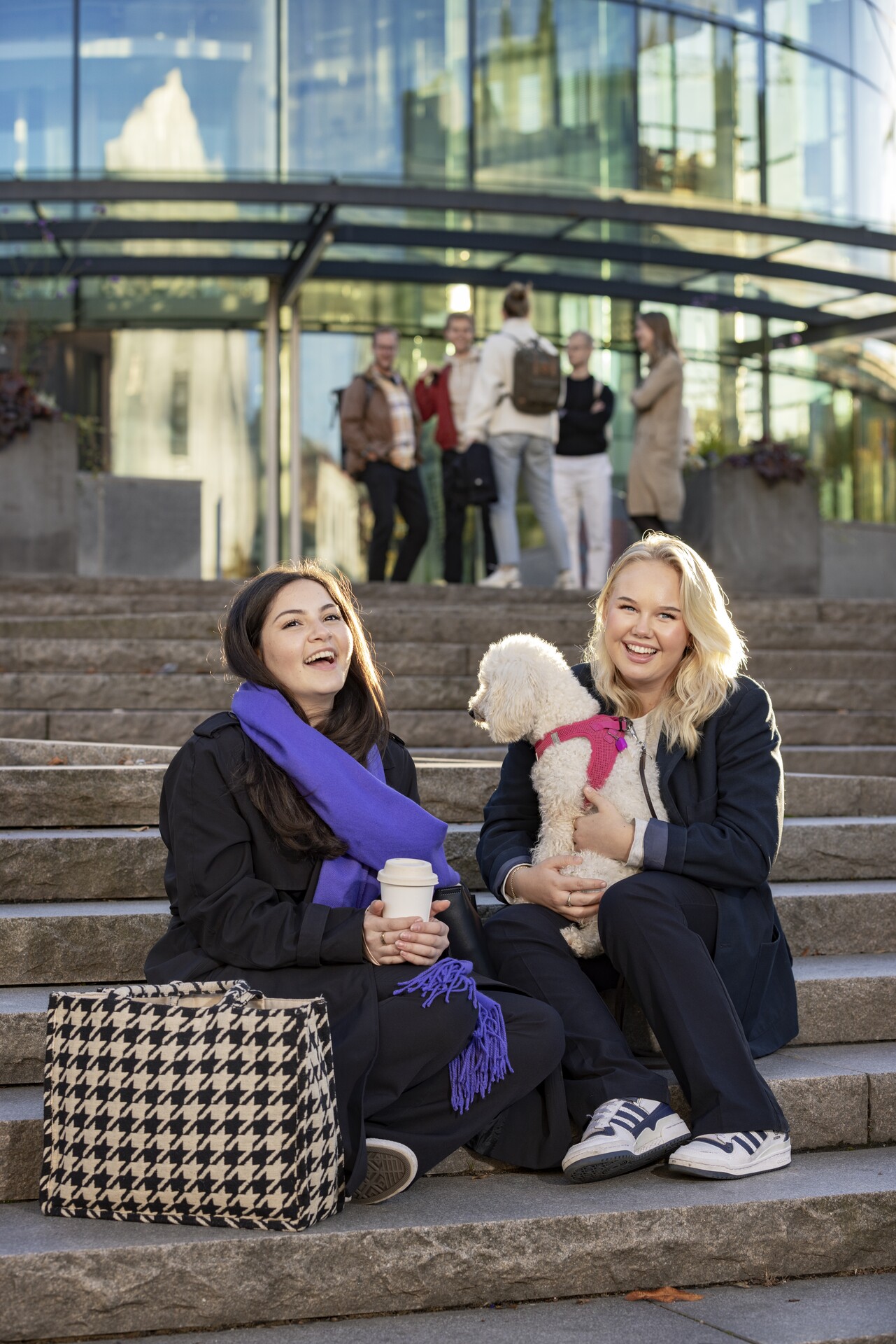 Två leende kvinnor sitter på en trappa utomhus framför en modern glasbyggnad. Den ena håller en takeaway-kaffemugg och har en lila halsduk, den andra håller en liten vit hund i rosa sele i knät. Bredvid dem står en stor tygväska med hundtandsmönster, och i bakgrunden syns en grupp människor som står och pratar.