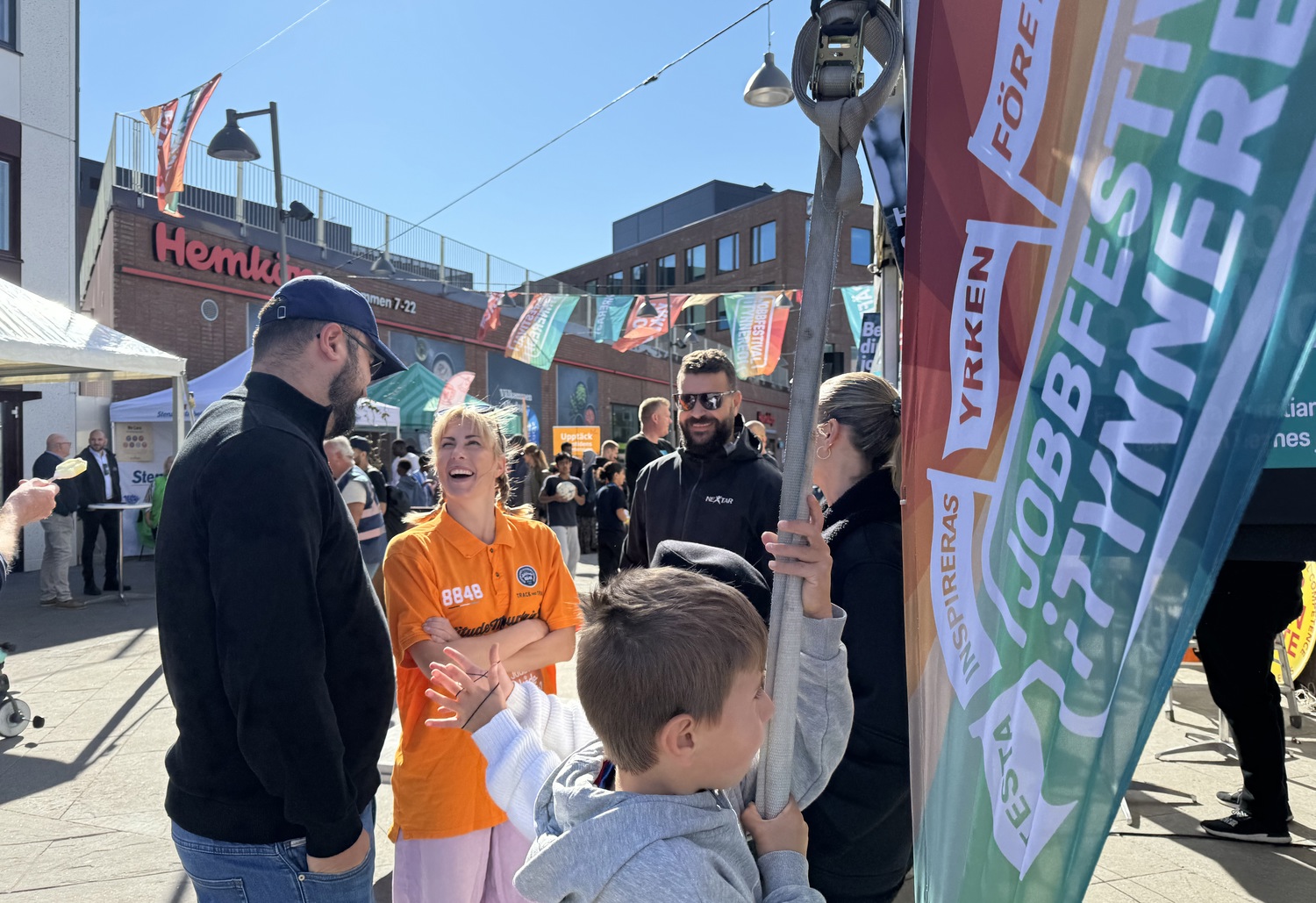 Människor möts och samtalar på Jobbfestivalen i Tynnered vid Opaltorget, med färgglada flaggor och aktiviteter i soligt väder.