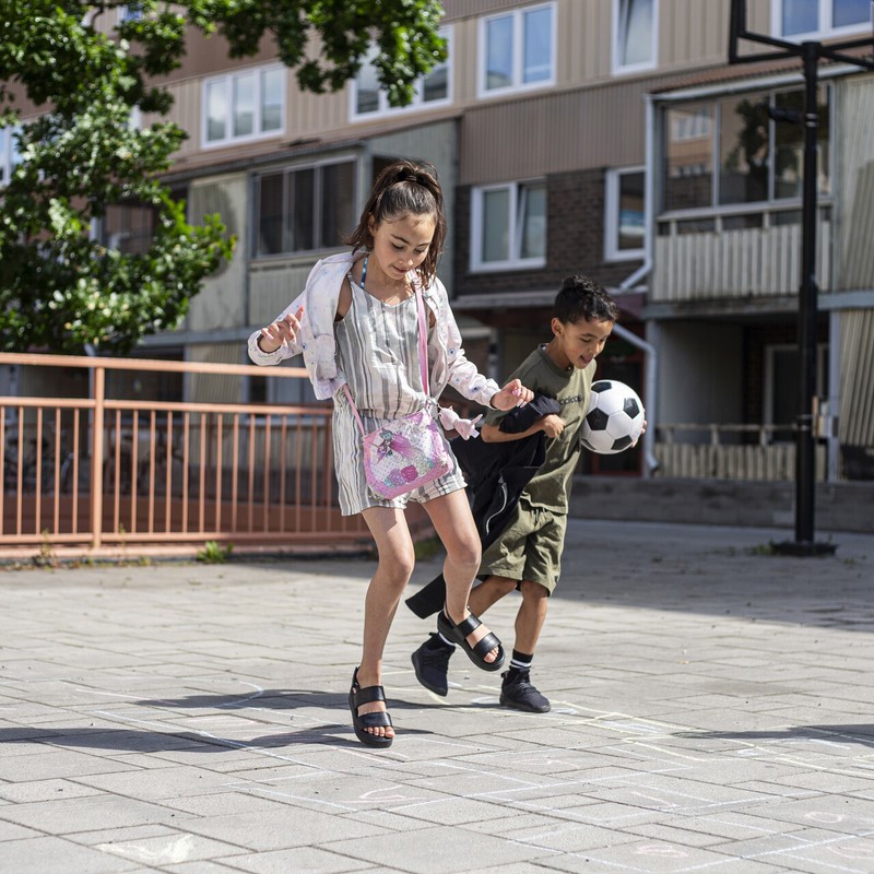 Två barn leker på en solig gård framför ett flerbostadshus; flickan hoppar på ett hopphagaritat med gatukritor medan pojken bredvid spelar fotbol.