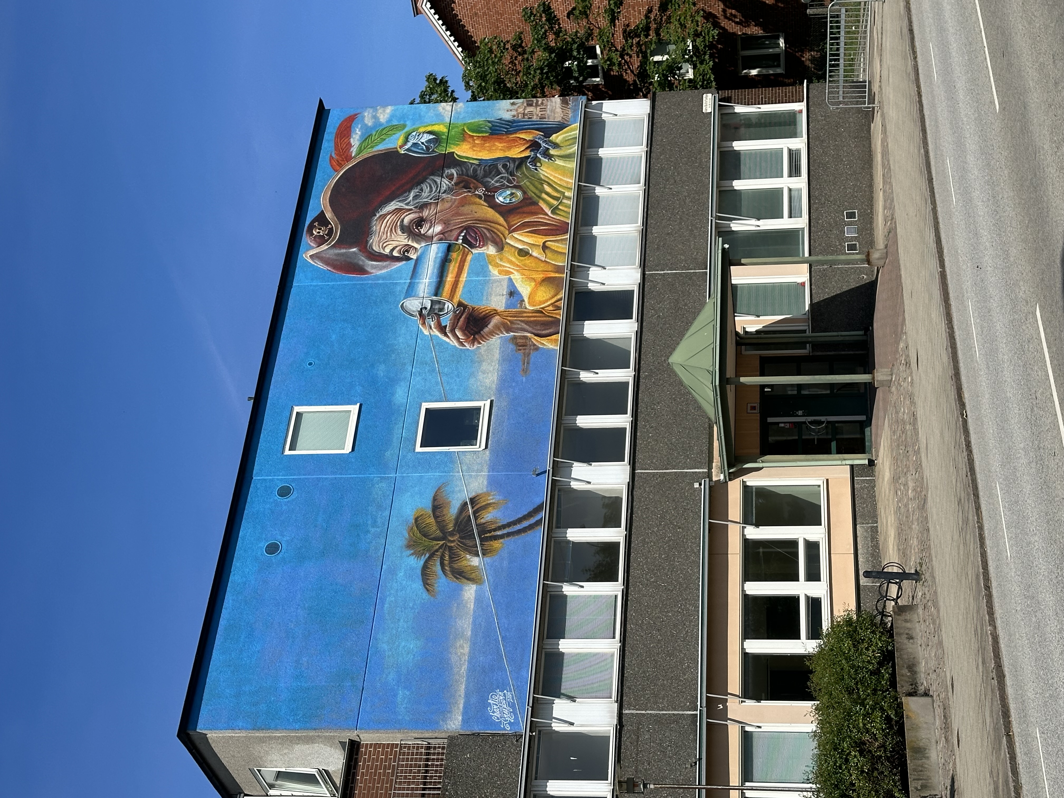 En byggnad med en stor väggmålning föreställande en färgglad pirat som håller i ett glas. Bakgrunden av målningen visar en blå himmel och en palm. Byggnaden har flera fönster och en grön entré. Gatan framför är tom och himlen är klarblå. Stämningen är livfull och konstnärlig.