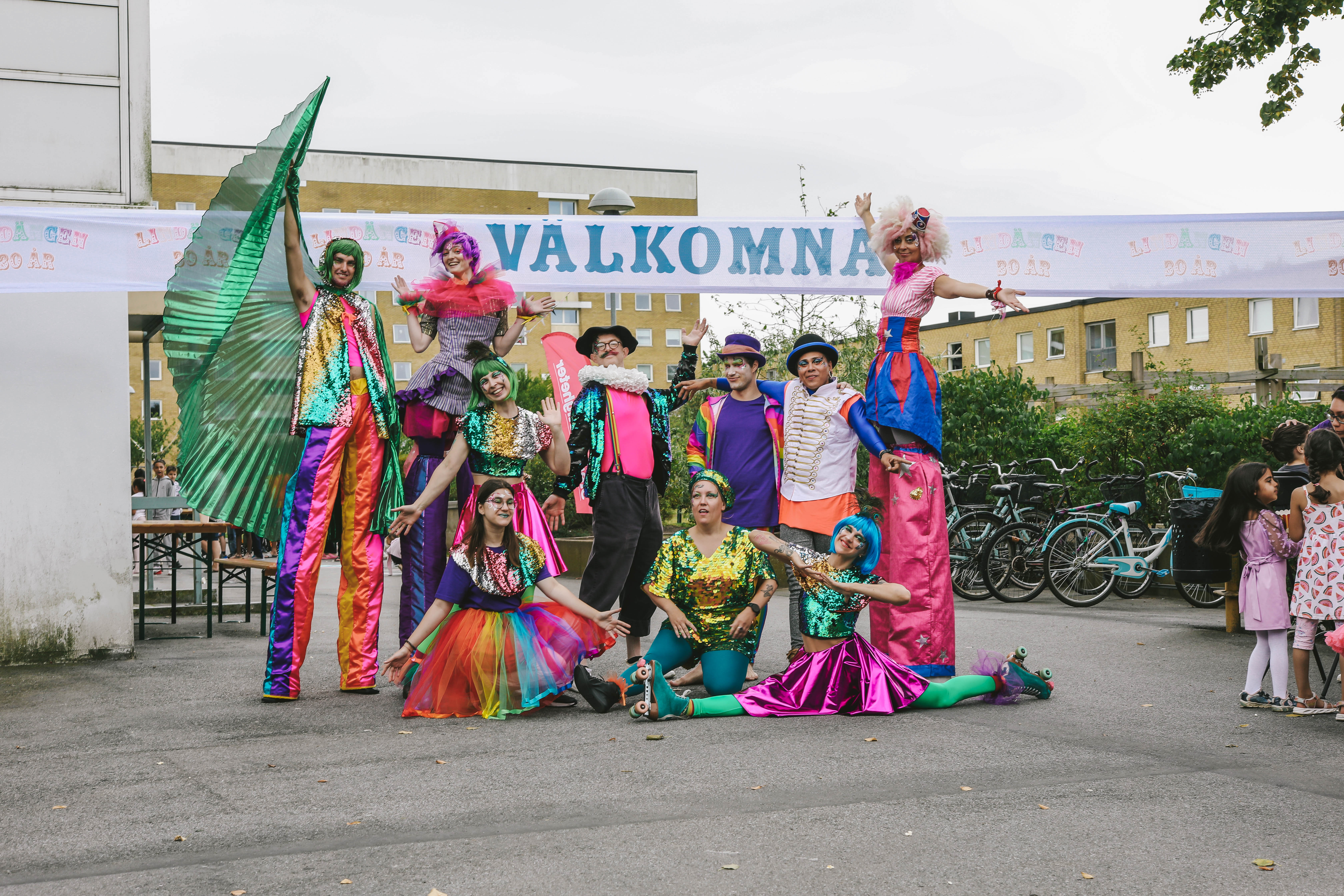 Cirkusgrupp i färgglada kläder poserar under en skylt med texten Välkomna