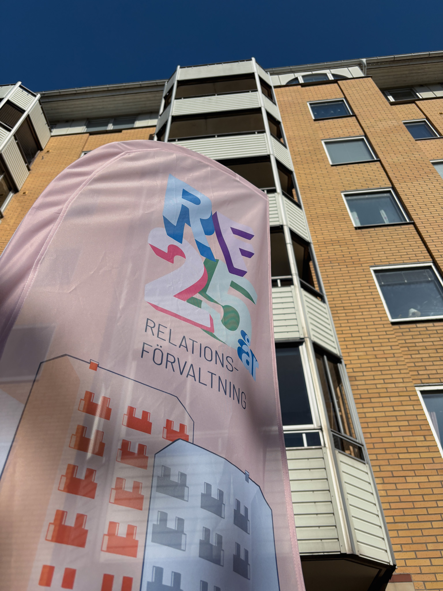  Vinklad närbild underifrån på en rosa flagga med texten ”RF 25 år – Relationsförvaltning” framför ett gulbrunt flerbostadshus mot klar blå himmel.
