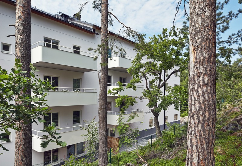 Ljus flerbostadsbyggnad med balkonger omgiven av höga tallar och grön skogsslänt under en blå himmel. Ängby Park Blackebergs gård Stockholm