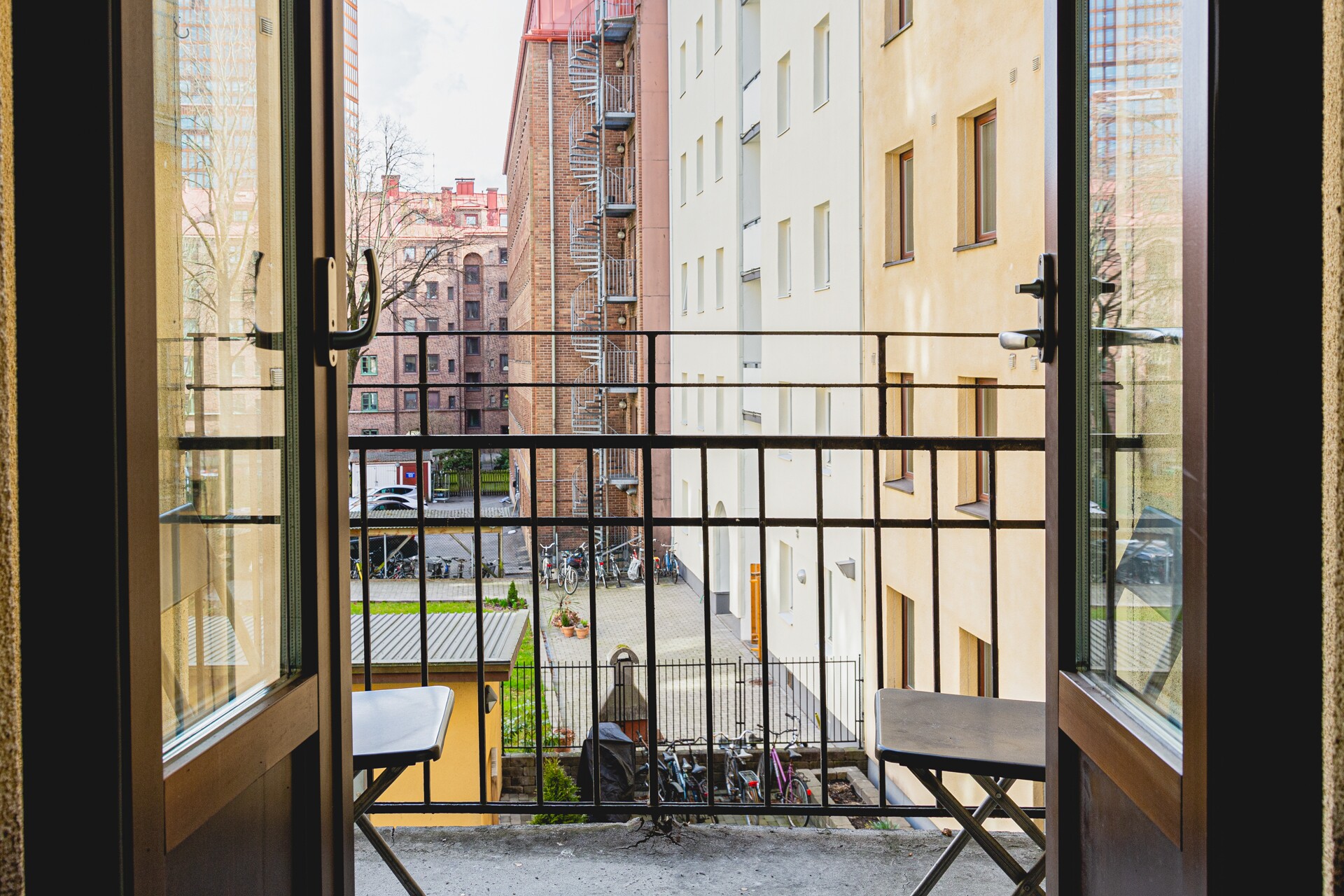 Liten balkong med två hopfällbara bord/stolar och svart räcke, sedd genom öppna glasdörrar, med utsikt mot innergård där cyklar står parkerade mellan ljusa och röda tegelhus med utvändig spiraltrappa.