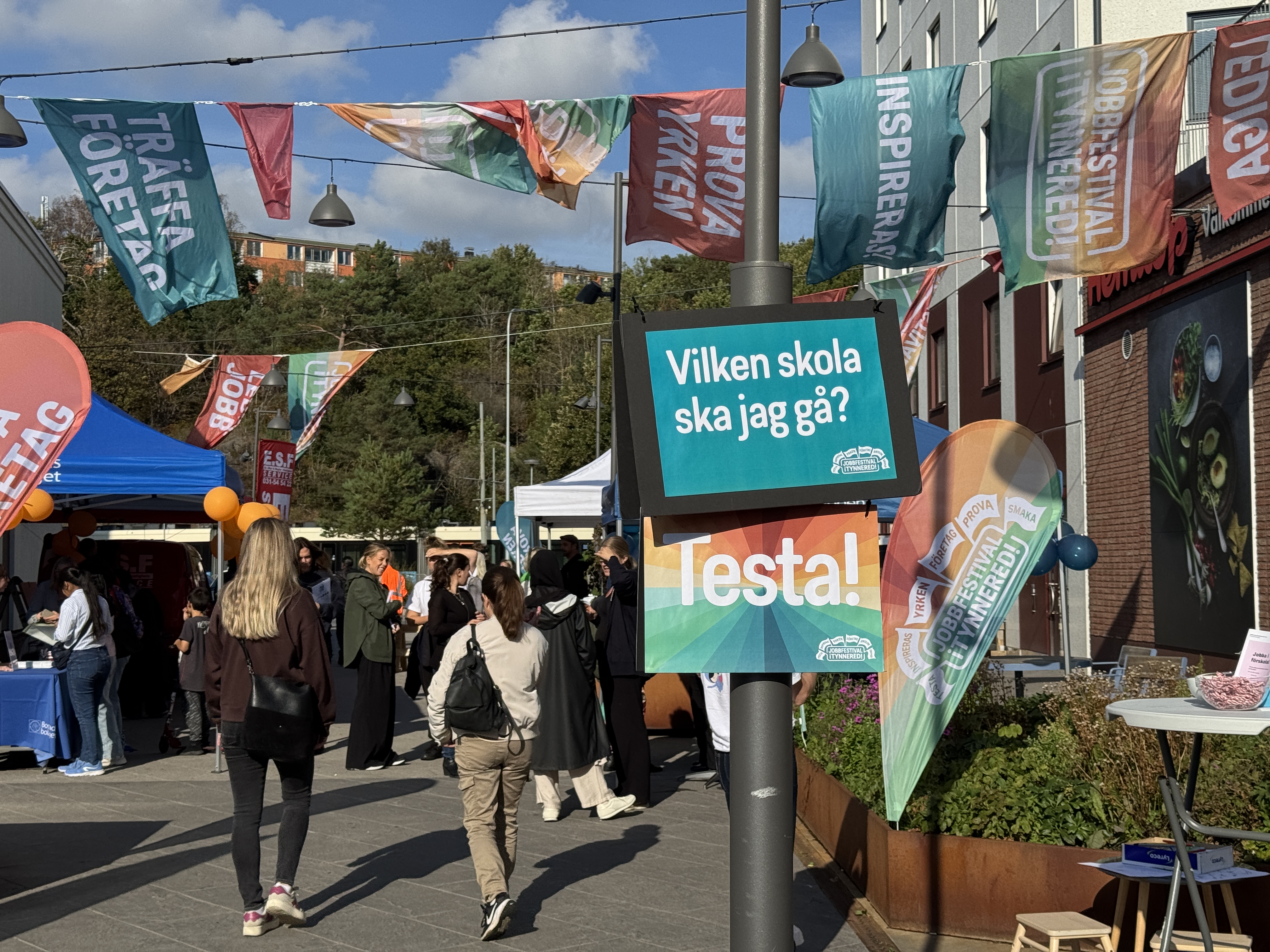 Utomhusmässa på ett torg med många besökare, färgglada tält och flaggor. I mitten sitter en skylt på en lyktstolpe med texten ”Vilken skola ska jag gå? Testa!”, och runtom syns fler banderoller med ord om att träffa företag och prova yrken. I bakgrunden skymtar träd och bostadshu