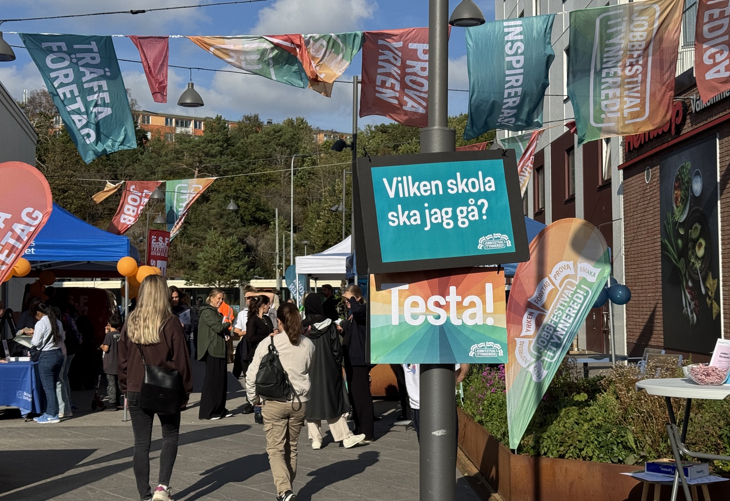 Utomhusmässa på ett torg med många besökare, färgglada tält och flaggor. I mitten sitter en skylt på en lyktstolpe med texten ”Vilken skola ska jag gå? Testa!”, och runtom syns fler banderoller med ord om att träffa företag och prova yrken. I bakgrunden skymtar träd och bostadshu