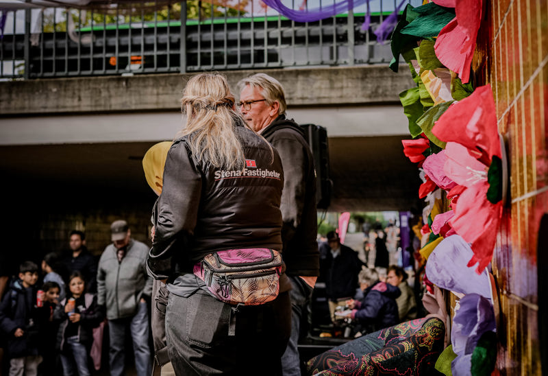 En grupp människor står utomhus vid ett evenemang. En person bär en jacka med text på ryggen. Färgglada dekorationer syns i förgrunden, och fler personer är samlade i bakgrunden. Stämningen verkar livlig och social.