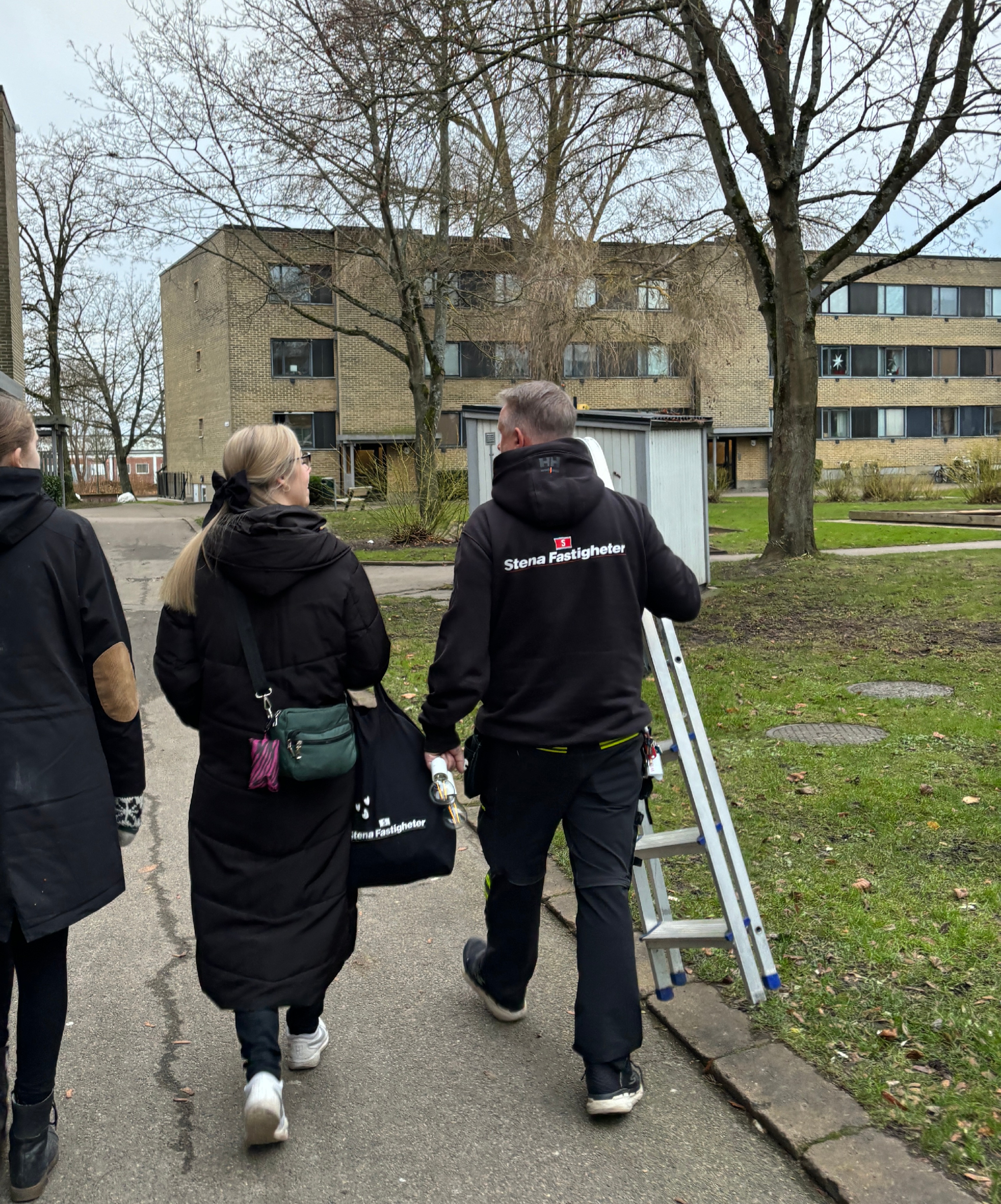 Tre personer går på en trottoar i ett bostadsområde. En av dem bär en jacka med texten "Stena Fastigheter" och håller i en hopfälld stege. De andra två är klädda i mörka jackor.