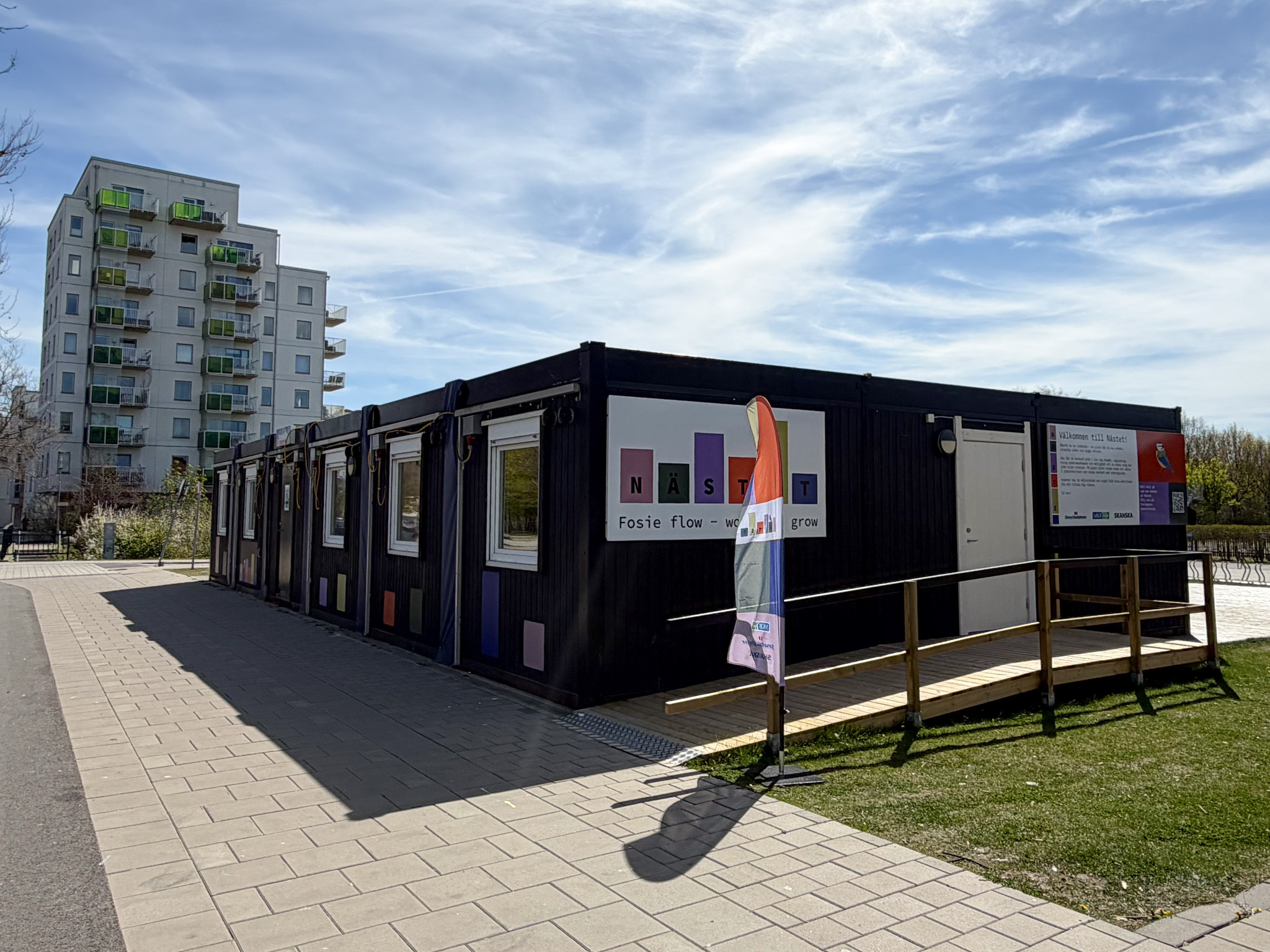 En låg, svart modulbyggnad med färgglada skyltar och en flagga utanför står längs en gång- och cykelväg. I bakgrunden syns ett högt bostadshus med balkonger och en blå vårhimmel med tunna moln.