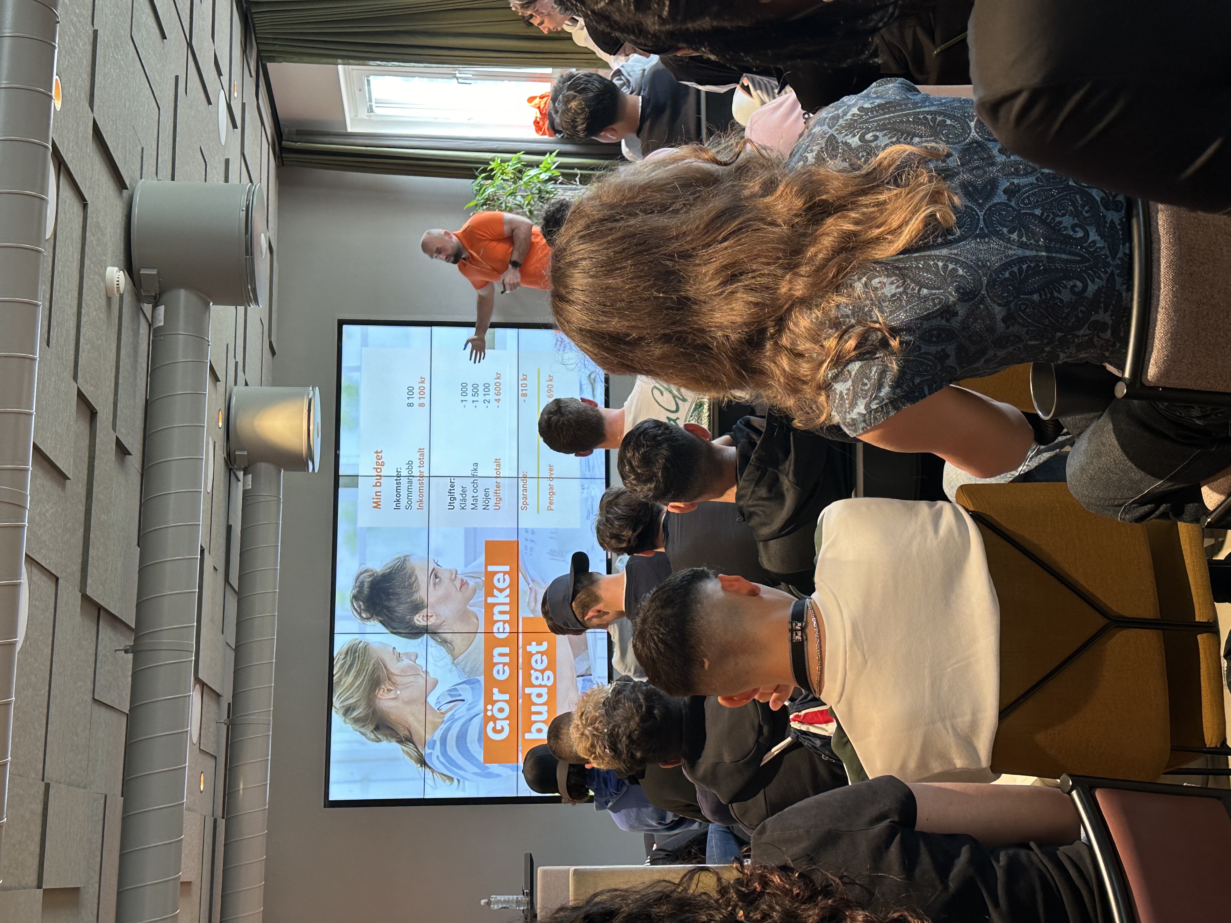 En föreläsare i orange tröja står framför en grupp människor och presenterar en bildskärm med texten "Gör en enkel budget". Deltagarna sitter vända mot skärmen och lyssnar uppmärksamt.