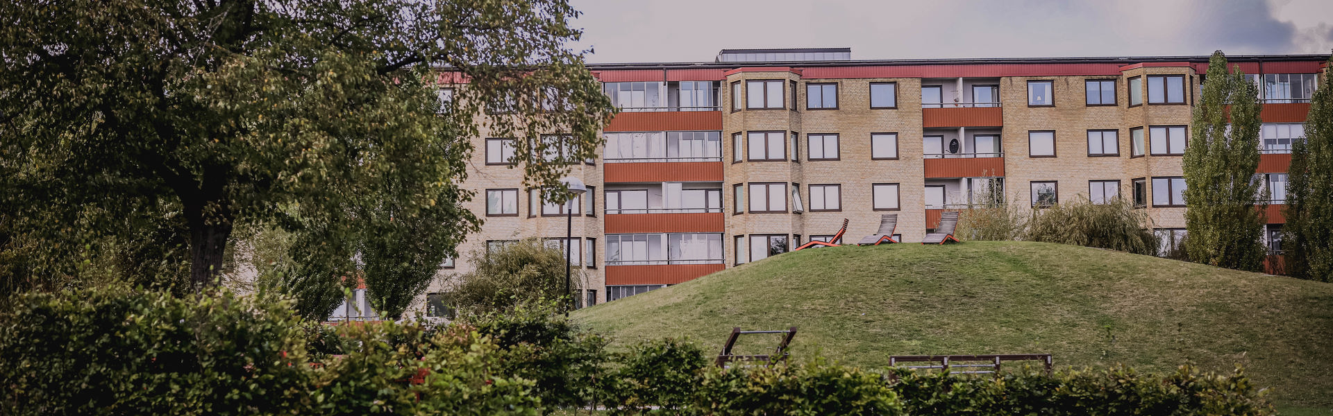Gönskande parkmiljö med röda stolar i förgrunden, i bakgrunden syns ett flerfamiljshus i gult tegel och röda balkongdetaljerGult tegel
