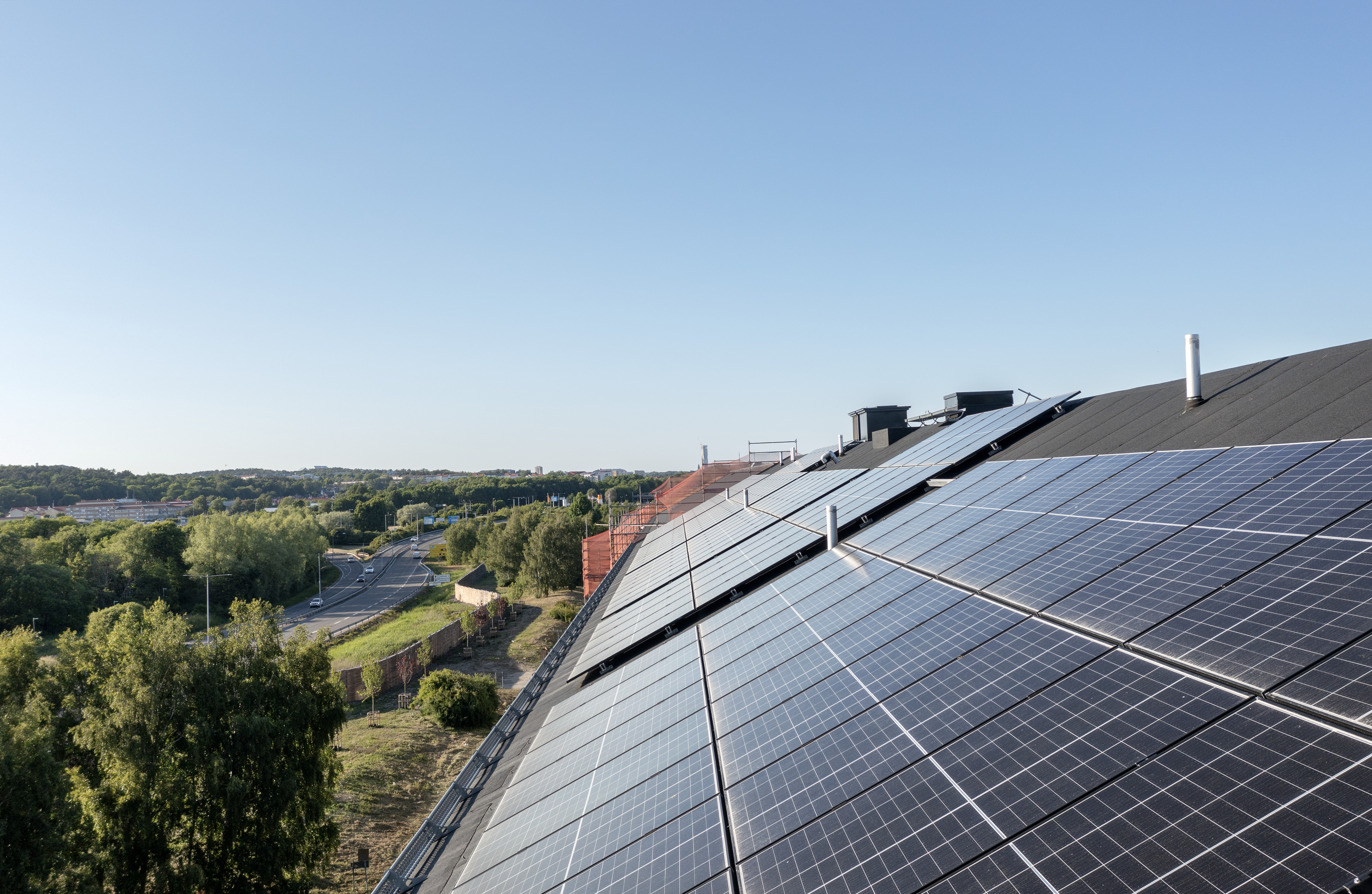 Solpaneler täcker ett snedtak på en byggnad i förgrunden, med grönskande träd, en väg med bilar och ett stadslandskap i bakgrunden under klar blå himmel.