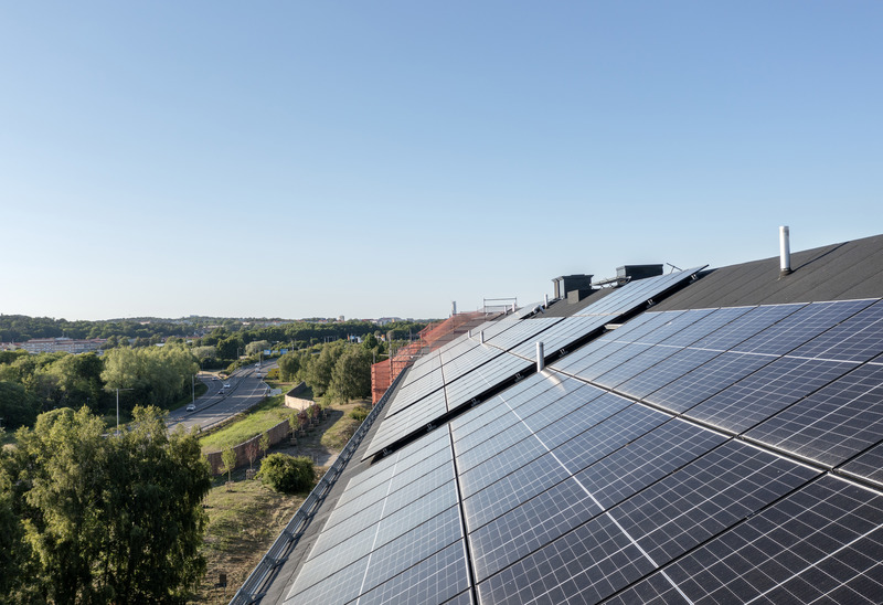 Solpaneler täcker ett snedtak på en byggnad i förgrunden, med grönskande träd, en väg med bilar och ett stadslandskap i bakgrunden under klar blå himmel.