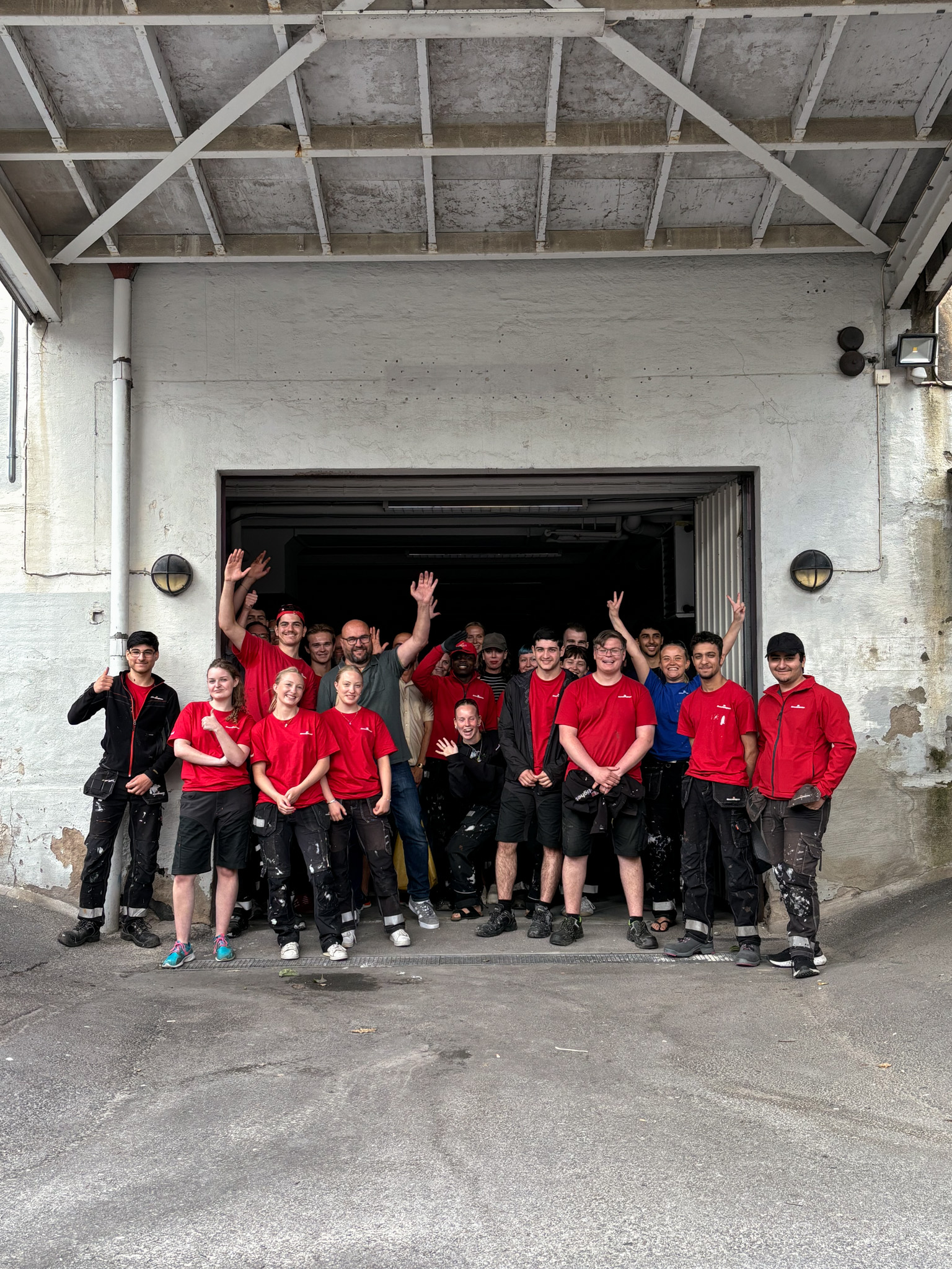 En grupp personer i arbetskläder, många i röda tröjor, står samlade framför en öppen garageport. Flera ler och vinkar mot kameran, vilket ger en glad och gemensam lagkänsla.