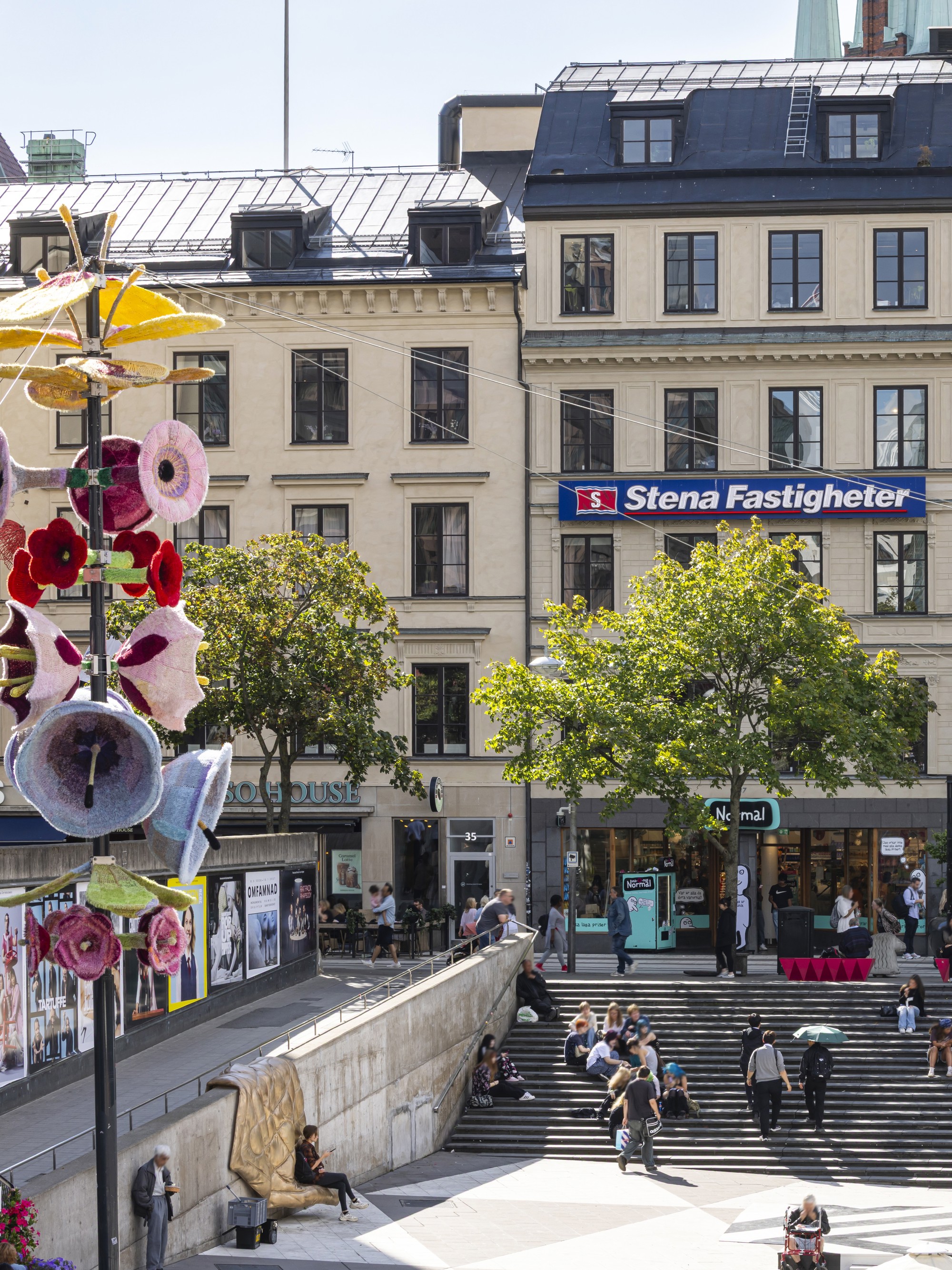 Vy över en stadsmiljö med äldre fastigheter, butikslokaler i gatuplan och en bred trappa där människor sitter och rör sig. På en fasad syns en skylt med texten Stena Fastigheter. I förgrunden finns träd, en upphöjd gångyta och färgstarka blomliknande utsmyckningar på stolpar.