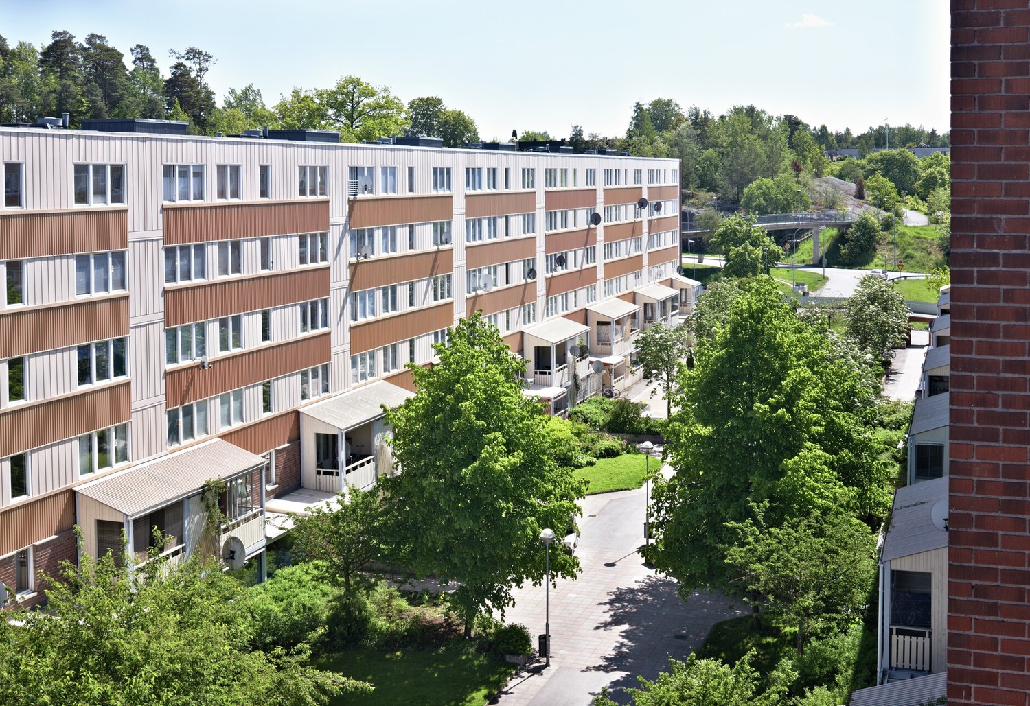 Lägenhetshus i Fisksätra, Stockholm en solig dag. Bakom huset syns grönska och träd. Framför huset finns gräsplättar, fler träd och lyktstolpar. Bilden är tagen ovanifrån från ett hus mittemot 