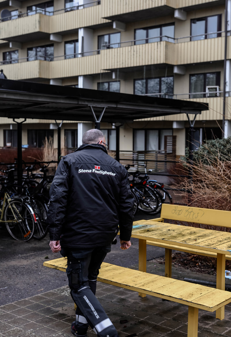 En person i en jacka med Stena Fastigheter-logga på ryggen står vid ett cykelskjul. I bakgrunden syns en flerfamiljsbyggnad med balkonger.