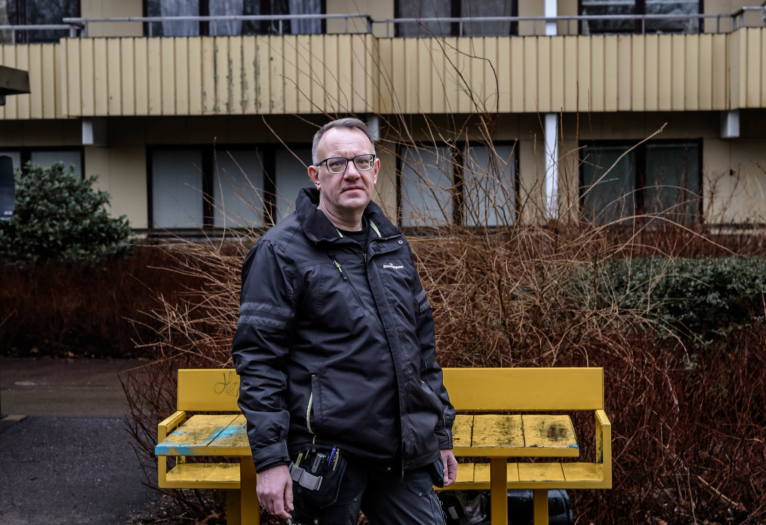 En person i arbetskläder står framför en gul bänk på en stenlagd yta. Bakom personen finns buskar och en flerfamiljsbyggnad med balkonger. Marken är fuktig, vilket tyder på nyligen regn.
