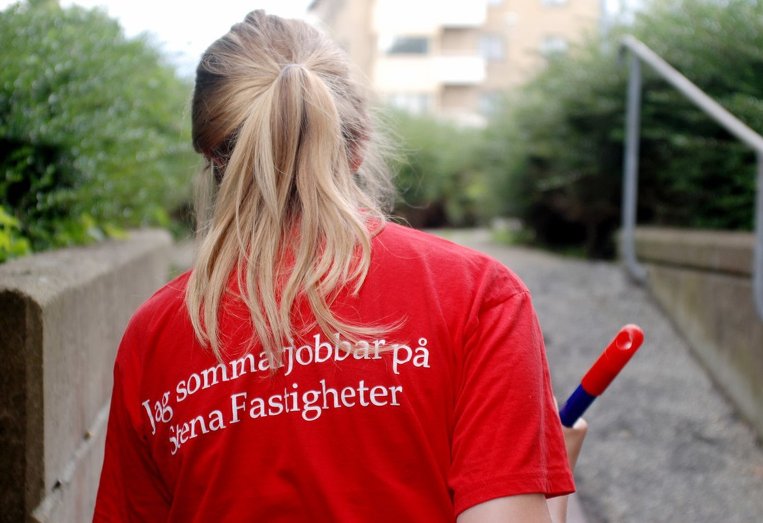Blond tjej fotad bakifrån. Hon bär en röd t-shirt med texten ´Jag sommarjobbar på Stena Fastigheter` på ryggen. 