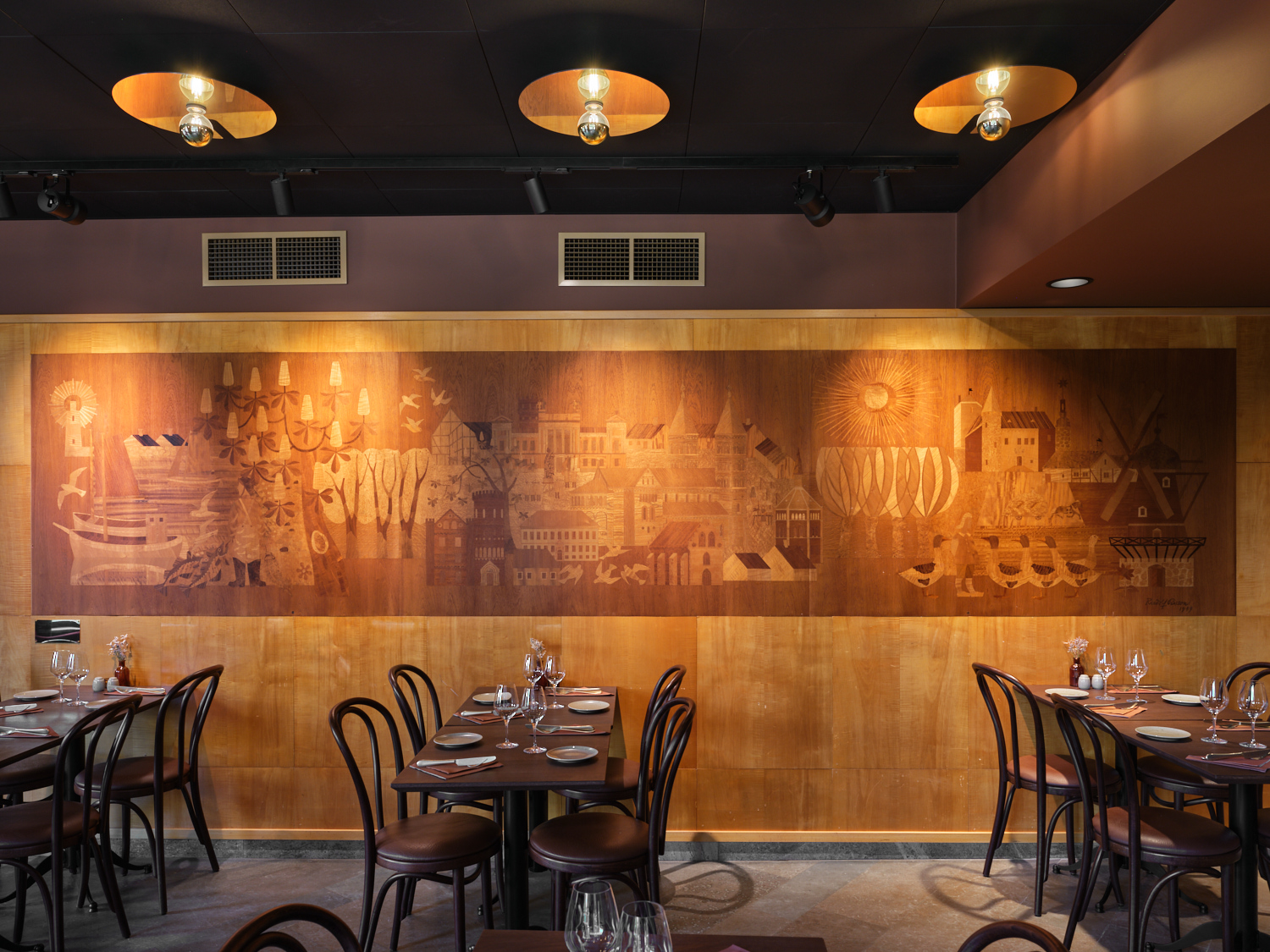 En varmrestaurangmiljö med mörka trästolar och dukade bord längs väggarna. Längs den bakre väggen löper ett stort, brunt konstverk i trä med inristade motiv av byggnader, träd och figurer, upplyst av spotlights från det mörka taket ovanför.