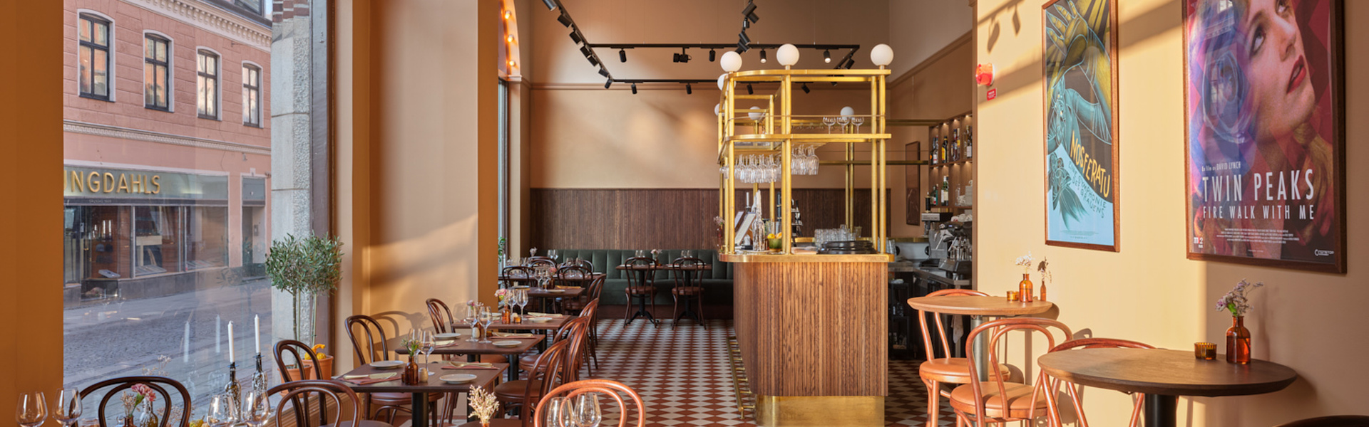 Ett ljust restaurangrum med rutigt rödvitt klinkergolv, höga fönster mot gatan och väggar i varma gula toner. Längs golvet står små dukade bord med böjträstolar, och längst in syns en bar med trä- och mässingsdetaljer, medan färgglada affischer pryder väggen till höger.