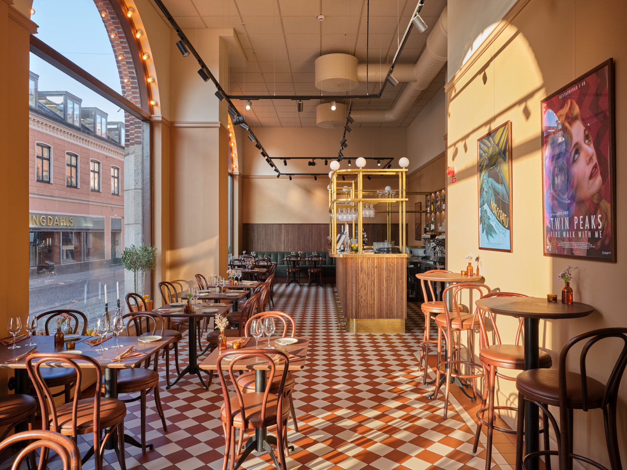 Ett ljust restaurangrum med rutigt rödvitt klinkergolv, höga fönster mot gatan och väggar i varma gula toner. Längs golvet står små dukade bord med böjträstolar, och längst in syns en bar med trä- och mässingsdetaljer, medan färgglada affischer pryder väggen till höger.