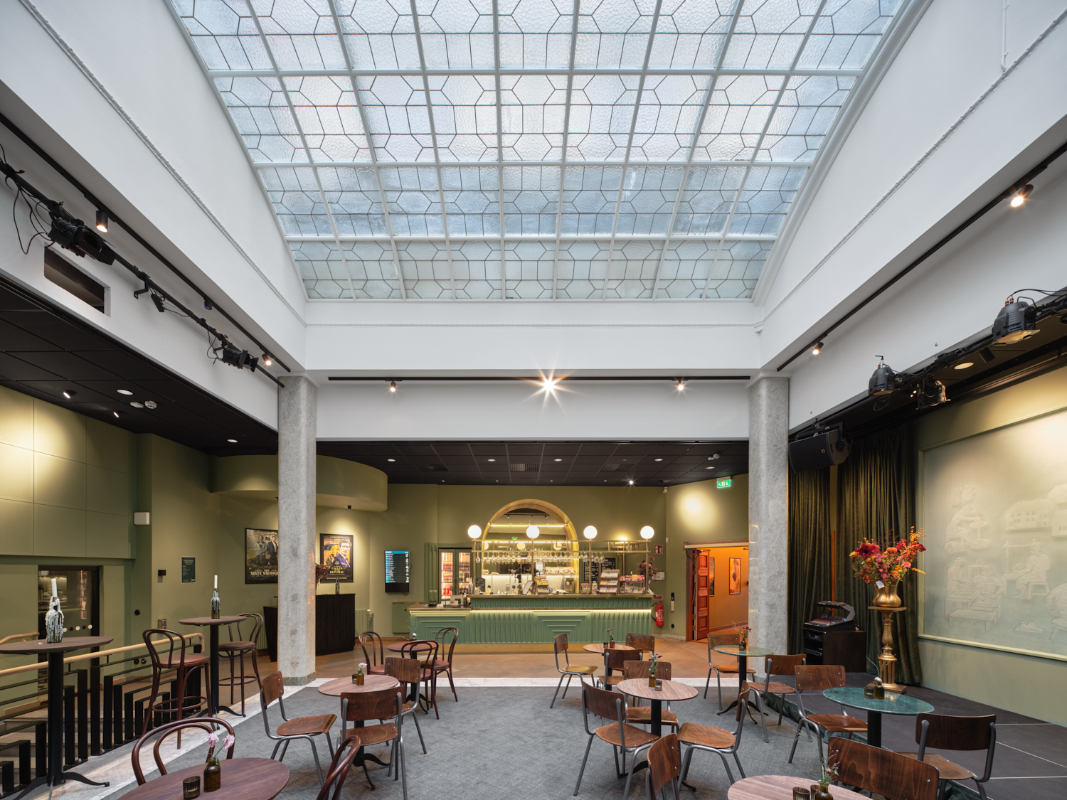 En rymlig foajé med högt glastak som släpper in ljus över ett öppet sällskapsutrymme med små runda bord och blandade trästolar. Längst bort syns en bar med grön front och väl upplyst serveringsdisk, omgiven av gröna väggar och scenutrustning, medan en hög vas med blommor står på ett sidobord till höger.