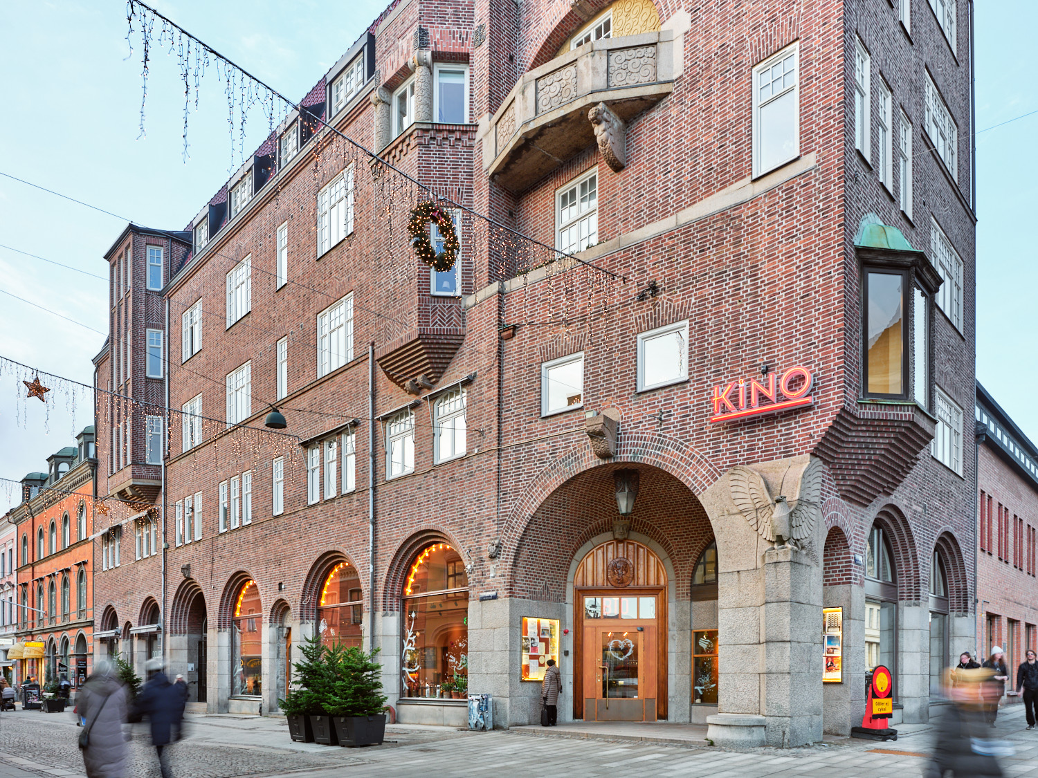 En exteriörbild av en tegelbyggnad i hörnläge med stora rundbågiga entréer i gatunivå och flera våningar ovanför. På fasaden sitter en neonskylt med texten ”KINO”, och framför huset rör sig människor längs trottoaren, medan julbelysning och en gran i kruka skapar stämning på gatan.