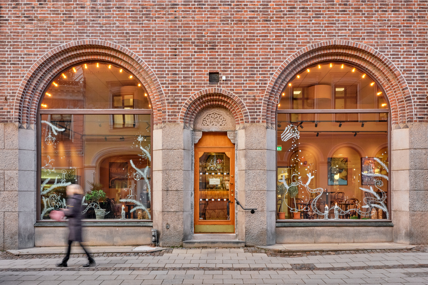 Exteriörbild av en tegelbyggnad med två stora rundbågiga skyltfönster och en trädörr i mitten, inramade av stenpelare. Genom fönstren syns den varmt upplysta restaurang- eller cafémiljön med bord och stolar, och på trottoaren utanför går en förbipasserande person i rörelse.