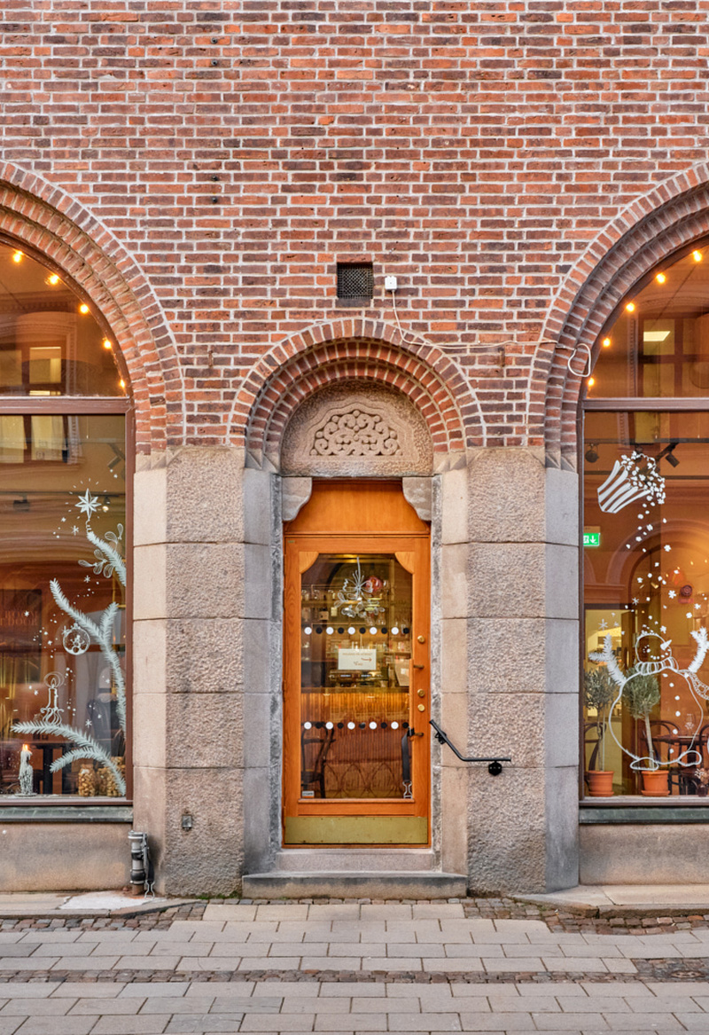 Exteriörbild av en tegelbyggnad med två stora rundbågiga skyltfönster och en trädörr i mitten, inramade av stenpelare. Genom fönstren syns den varmt upplysta restaurang- eller cafémiljön med bord och stolar, och på trottoaren utanför går en förbipasserande person i rörelse.