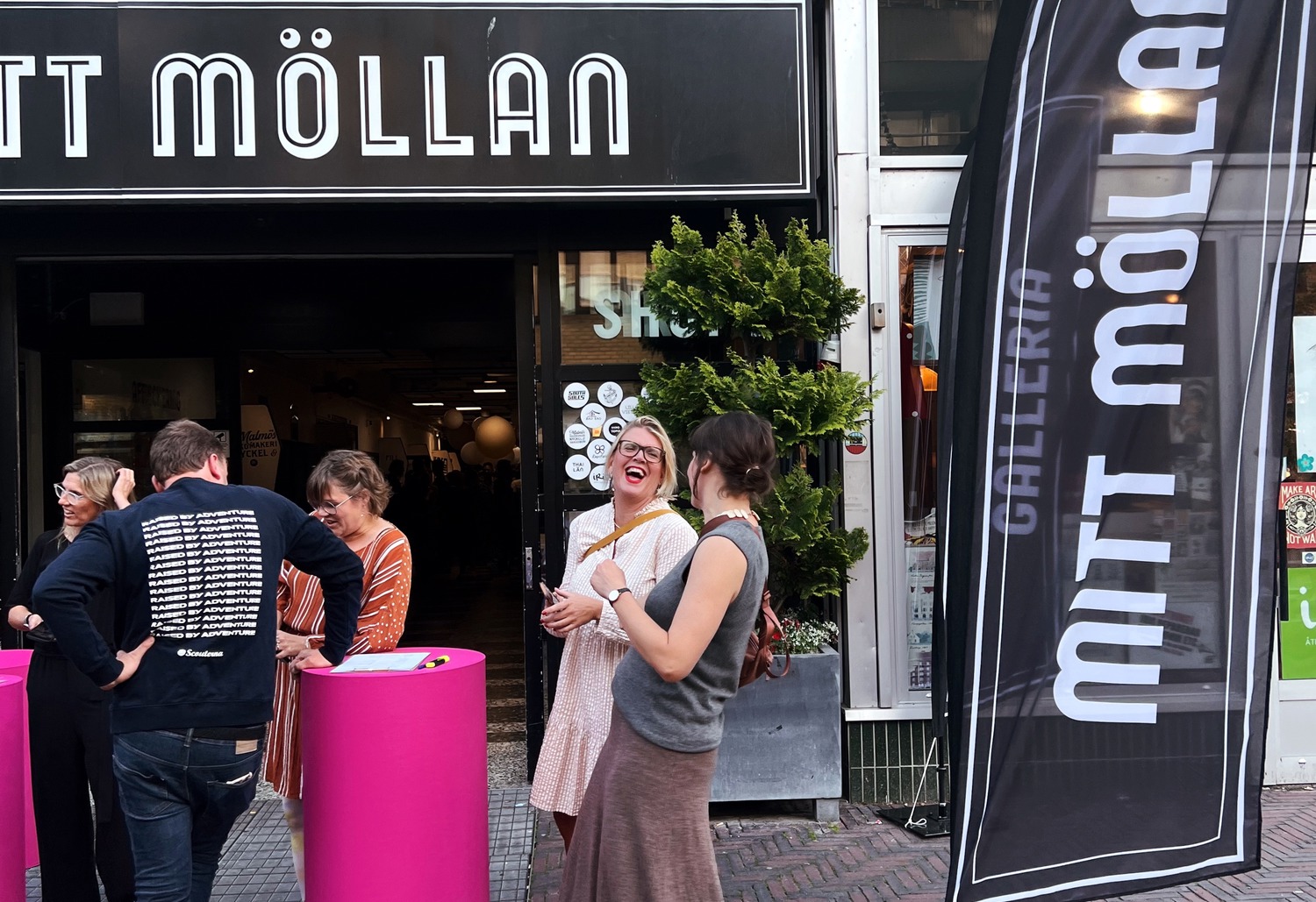 Alt-text: "Folk står och samtalar utanför entrén till Mitt Möllan. Framför ingången finns rosa ståbord och en svart flagga med texten 'Gallerian Mitt Möllan'. På fasaden ovanför syns vita masker med olika ansiktsuttryck.
