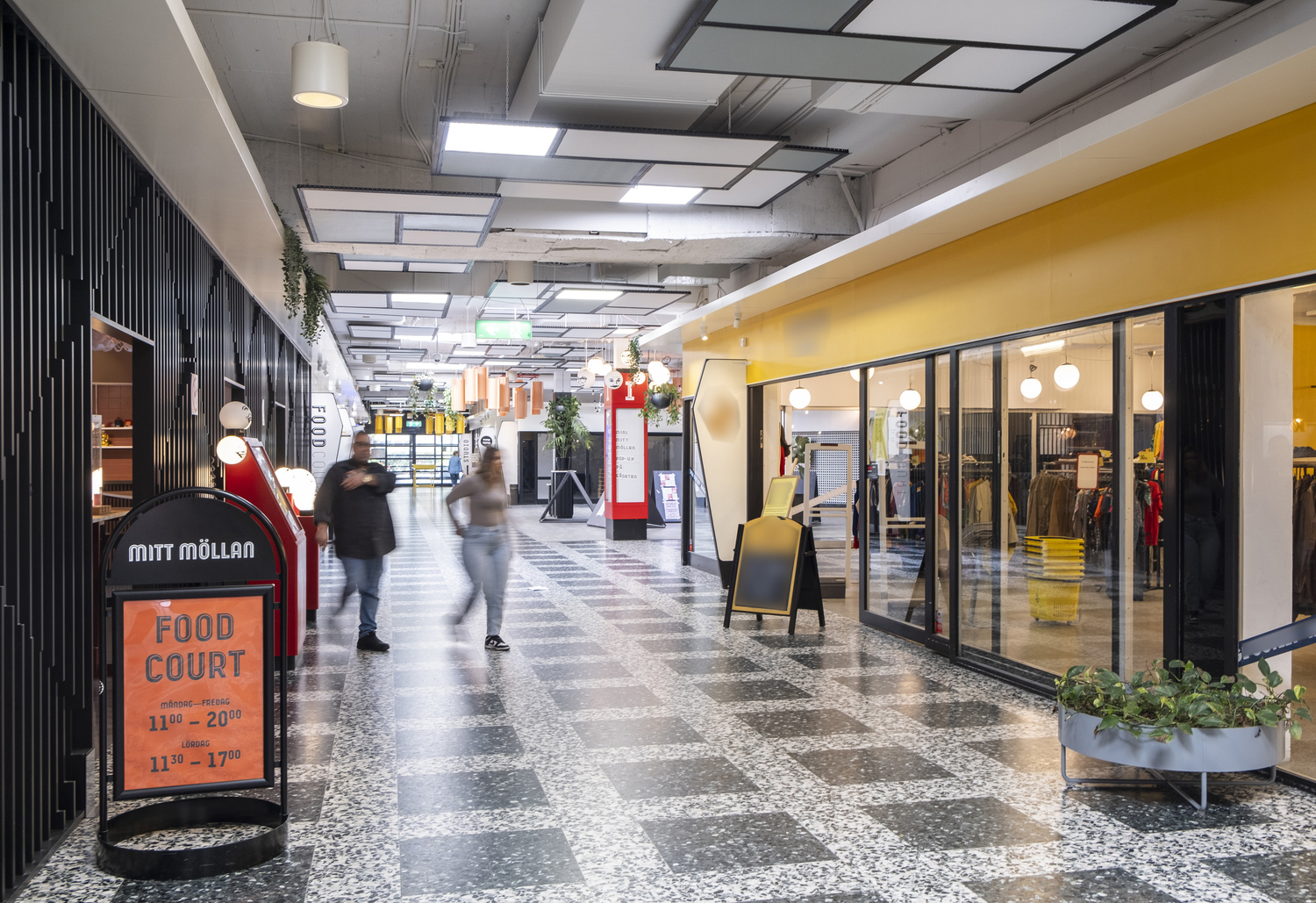 Inomhusbild av en modern galleria med människor som går förbi butiker och restauranger. Till vänster syns en skylt med texten 'Mitt Möllan Food Court'. Golvet har ett rutmönster och lokalen är ljus med stora fönster och taklampor