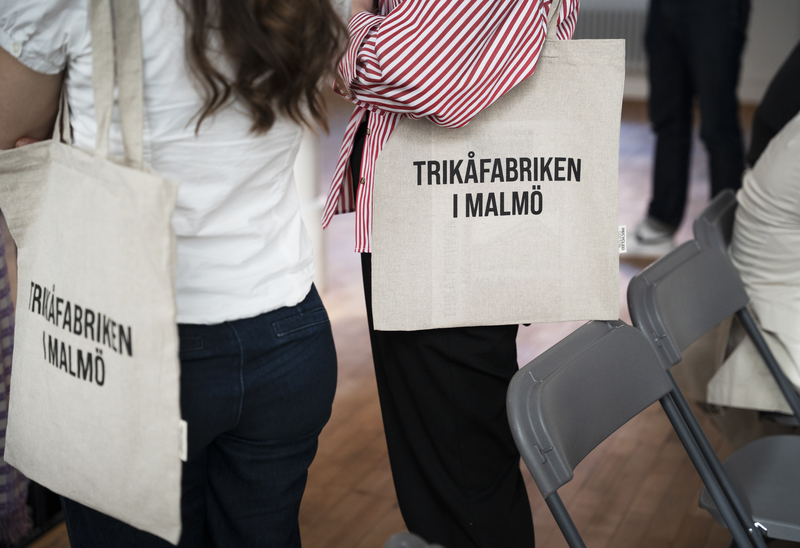 Två personer står bredvid varandra med tygkassar över axeln, båda med texten "TRIKÅFABRIKEN I MALMÖ" tryckt på väskan. De står i ett rum med hopfällbara stolar och trägolv.