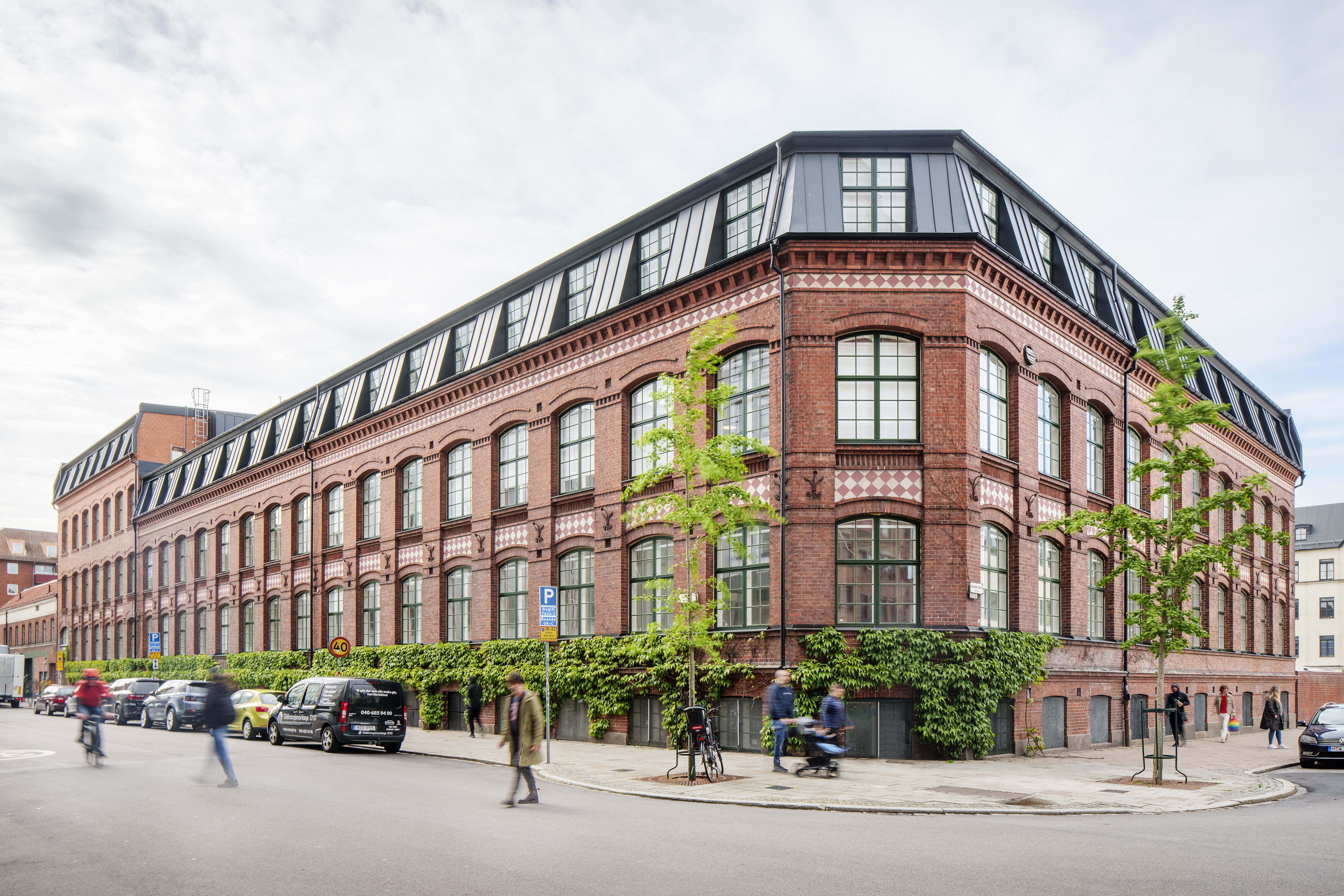Ett stort hörnhus i rött tegel med stora spröjsade fönster och svart takvåning, omgivet av träd, parkerade bilar och förbipasserande cyklister och fotgängare i en stadsmiljö.
