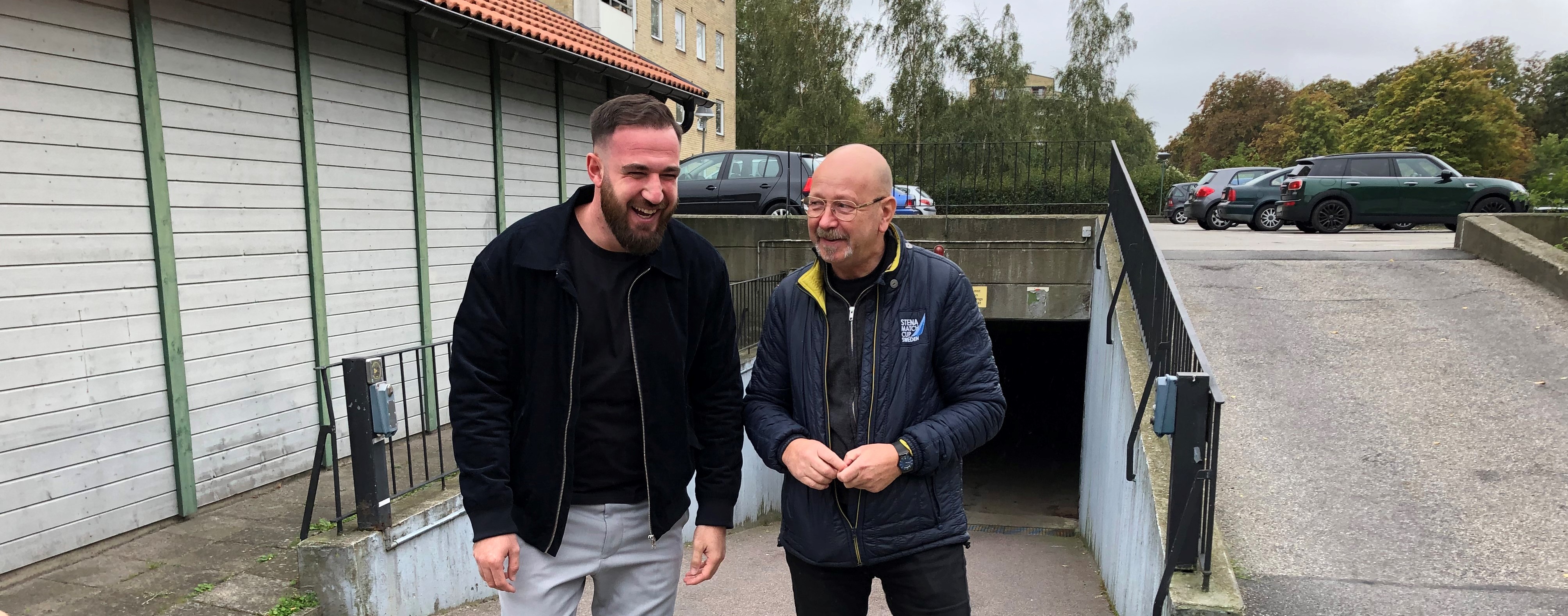 Två leende män promenerar tillsammans utomhus. Den ena mannen har kort hår och skägg, och bär en mörk jacka och ljusa byxor. Den andra mannen är skallig och bär glasögon, en mörk jacka och jeans. De går bredvid en undergång med en ramp och räcken. I bakgrunden syns parkerade bilar och träd.