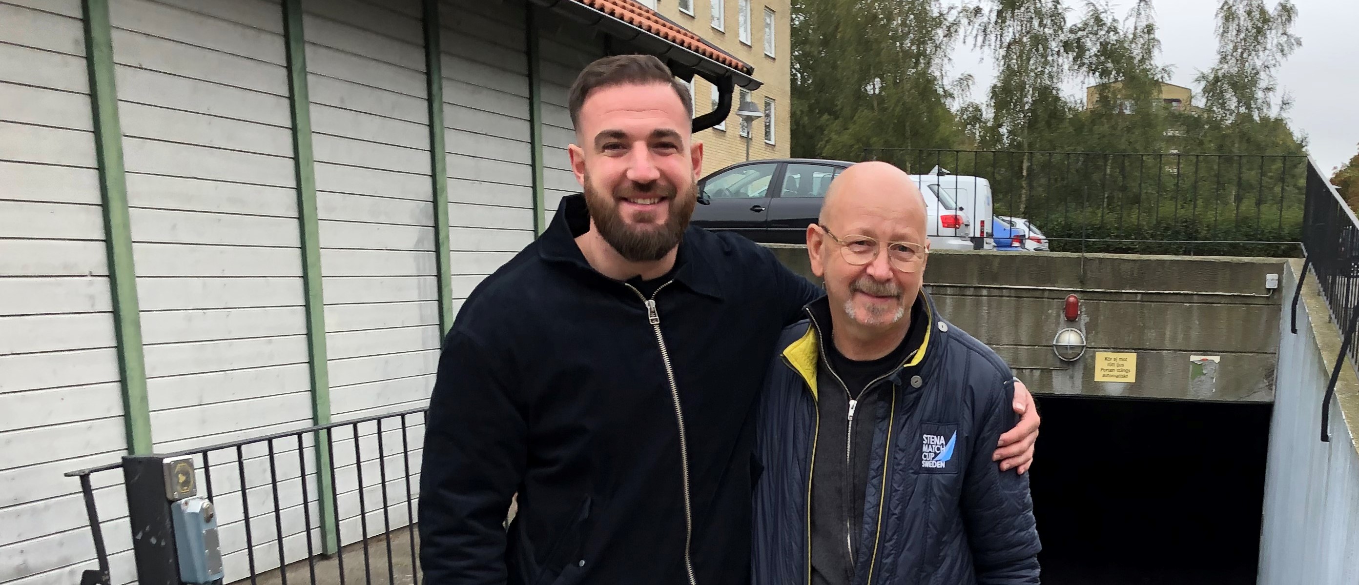 Två leende män står nära varandra utomhus. Den ena mannen har kort hår och skägg, och bär en mörk jacka. Den andra mannen är skallig, bär glasögon och en mörk jacka med en gul insida. De står vid en undergång med en ramp och räcken. I bakgrunden syns en parkerad bil och träd.