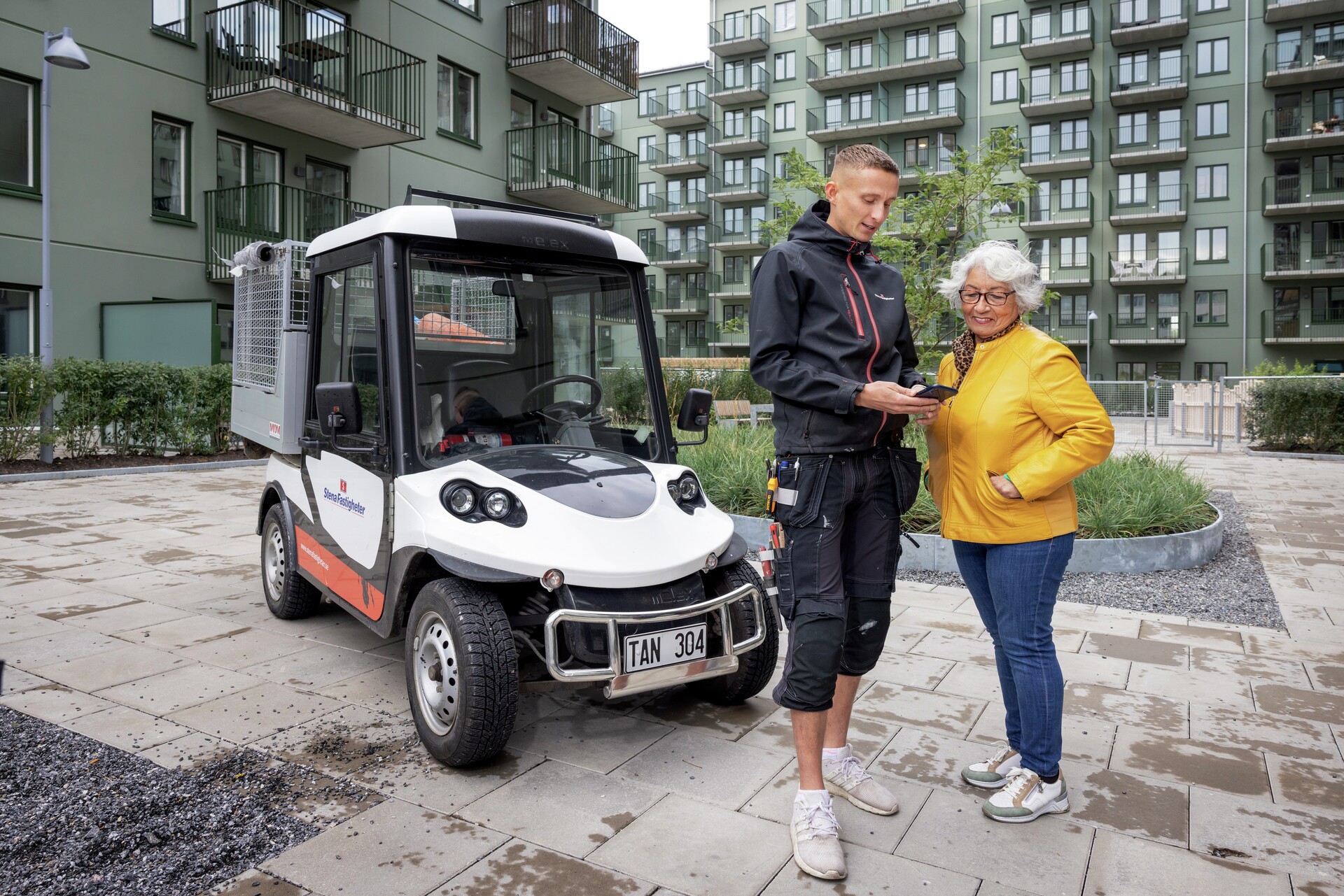 En yngre person i arbetskläder står bredvid en äldre kvinna i gul jacka på en gård mellan flerbostadshus. De tittar tillsammans på en mobiltelefon. Bredvid dem står ett litet vitt servicefordon parkerat på den stenlagda gången, med moderna lägenhetsbyggnader i bakgrunden.