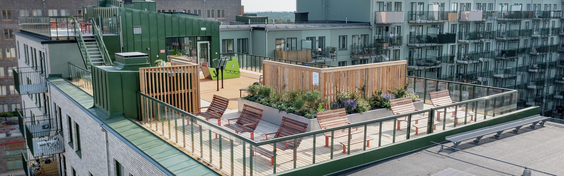 Ekebäckshöjd, Göteborg. En modern bostadsbyggnad med en stor takterrass syns ovanifrån, med trädeck, solstolar, bänkar och planteringar omgivna av glasräcken, medan flerbostadshus och solpaneler på ett intilliggande tak syns i bakgrunden under en klar himmel.