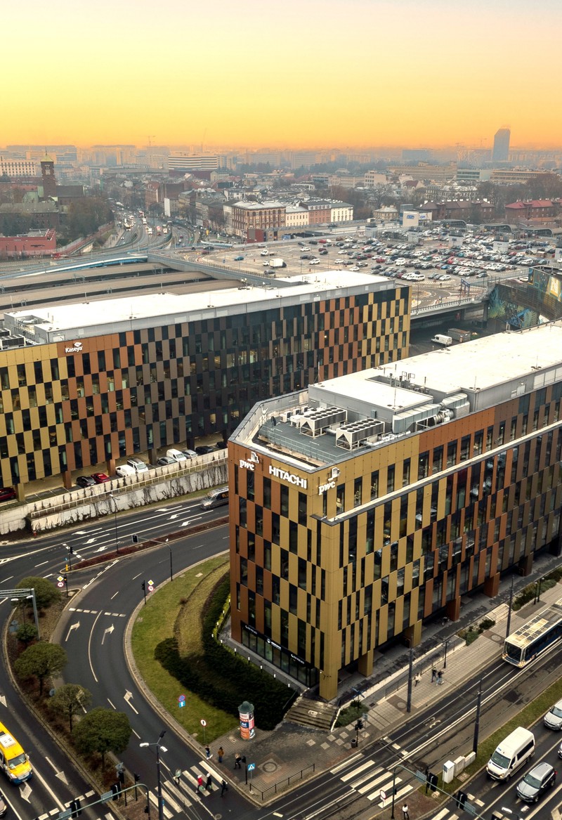 Aerial view of two modern office buildings with golden and dark panel facades, located along curved roads in a busy urban area. The scene includes multiple cars and buses, with a cityscape in the background under a warm, orange sunset sky.